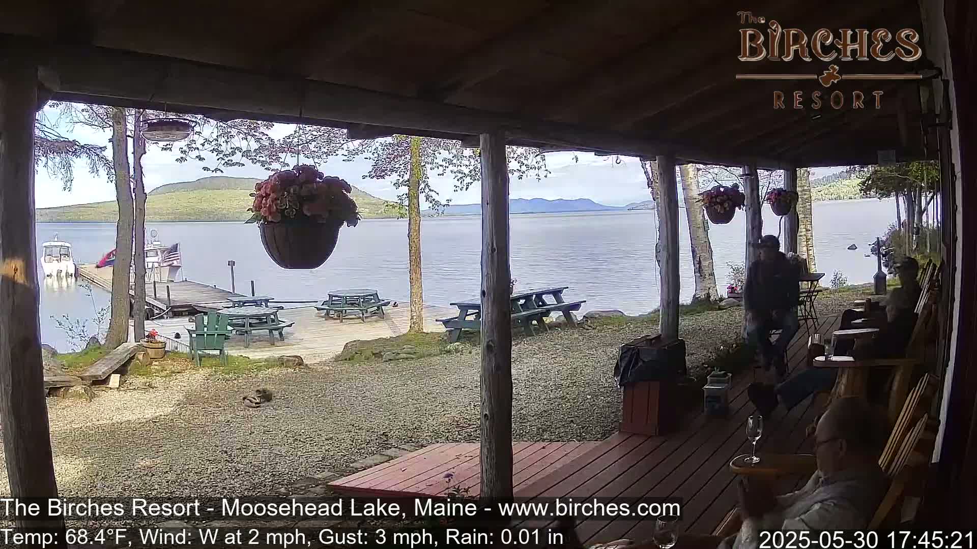 A lakeside porch with picnic tables and people sitting in wooden chairs, overlooking a calm lake with boats and mountains in the background under partly cloudy skies; light rain is falling.