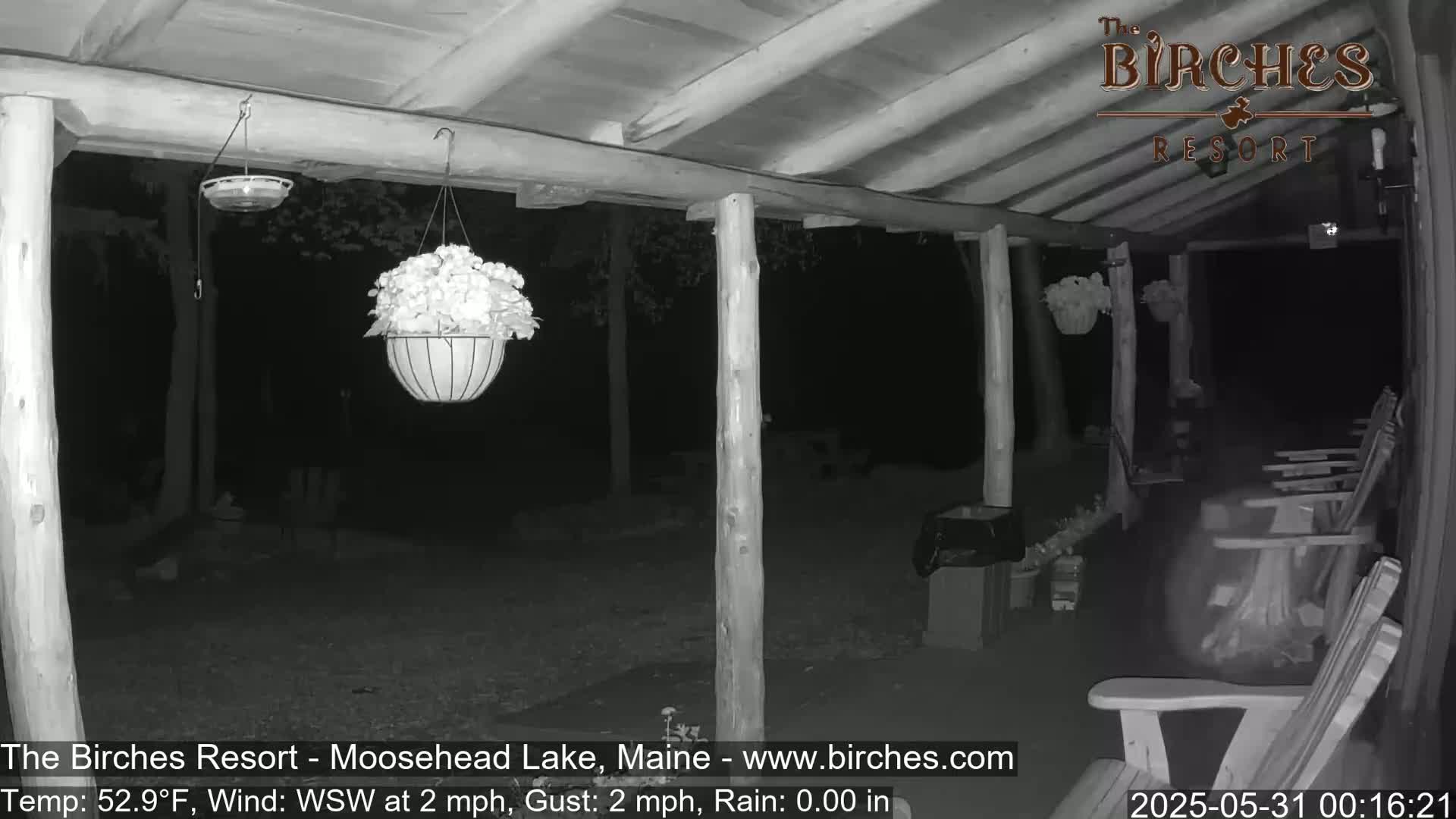 A nighttime, low-light view of a covered porch with hanging flower baskets and several empty rocking chairs, under partly cloudy skies.