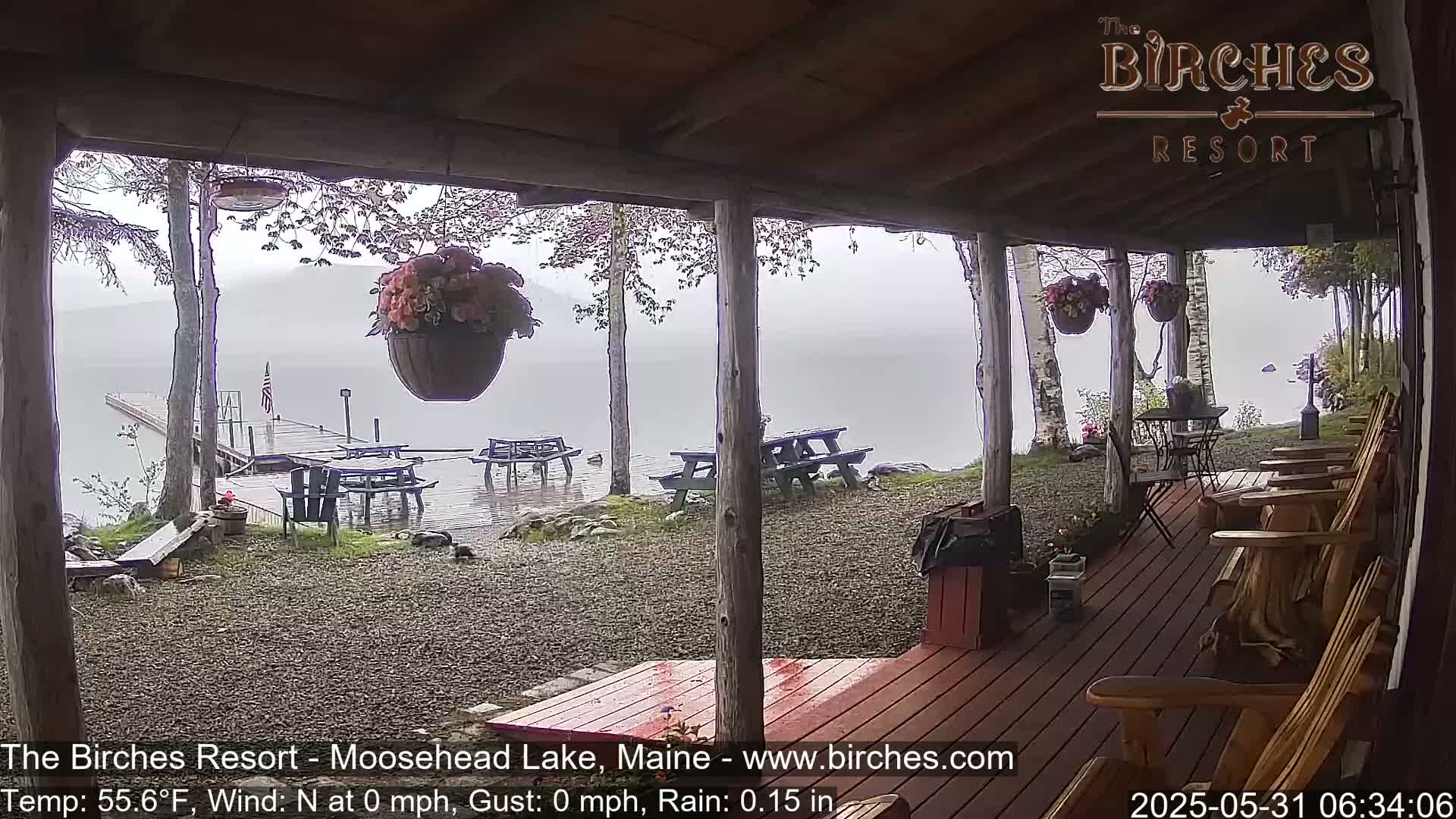 A rain-soaked porch overlooks a lake and dock, with picnic tables and Adirondack chairs scattered on the grounds.