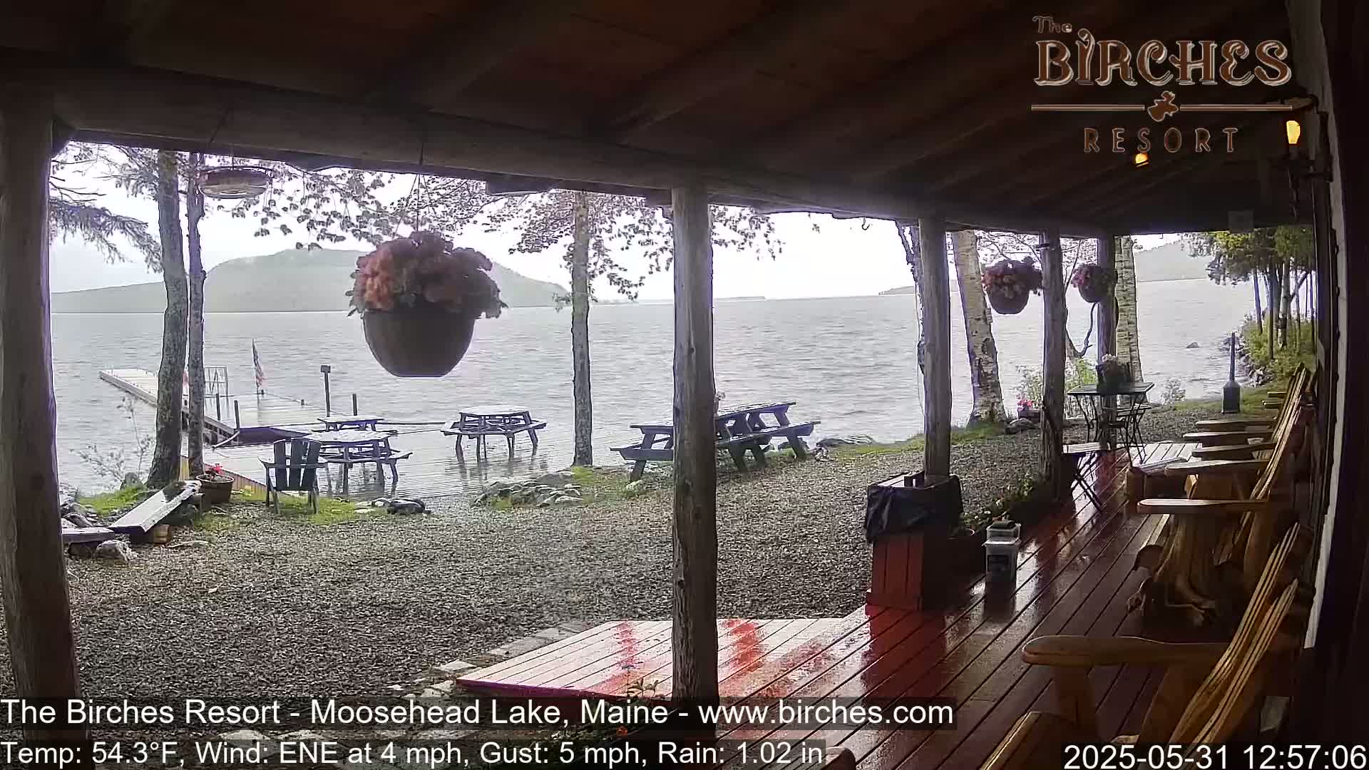 A rain-soaked porch overlooks a lake with picnic tables and Adirondack chairs on the ground, and a small island in the distance.