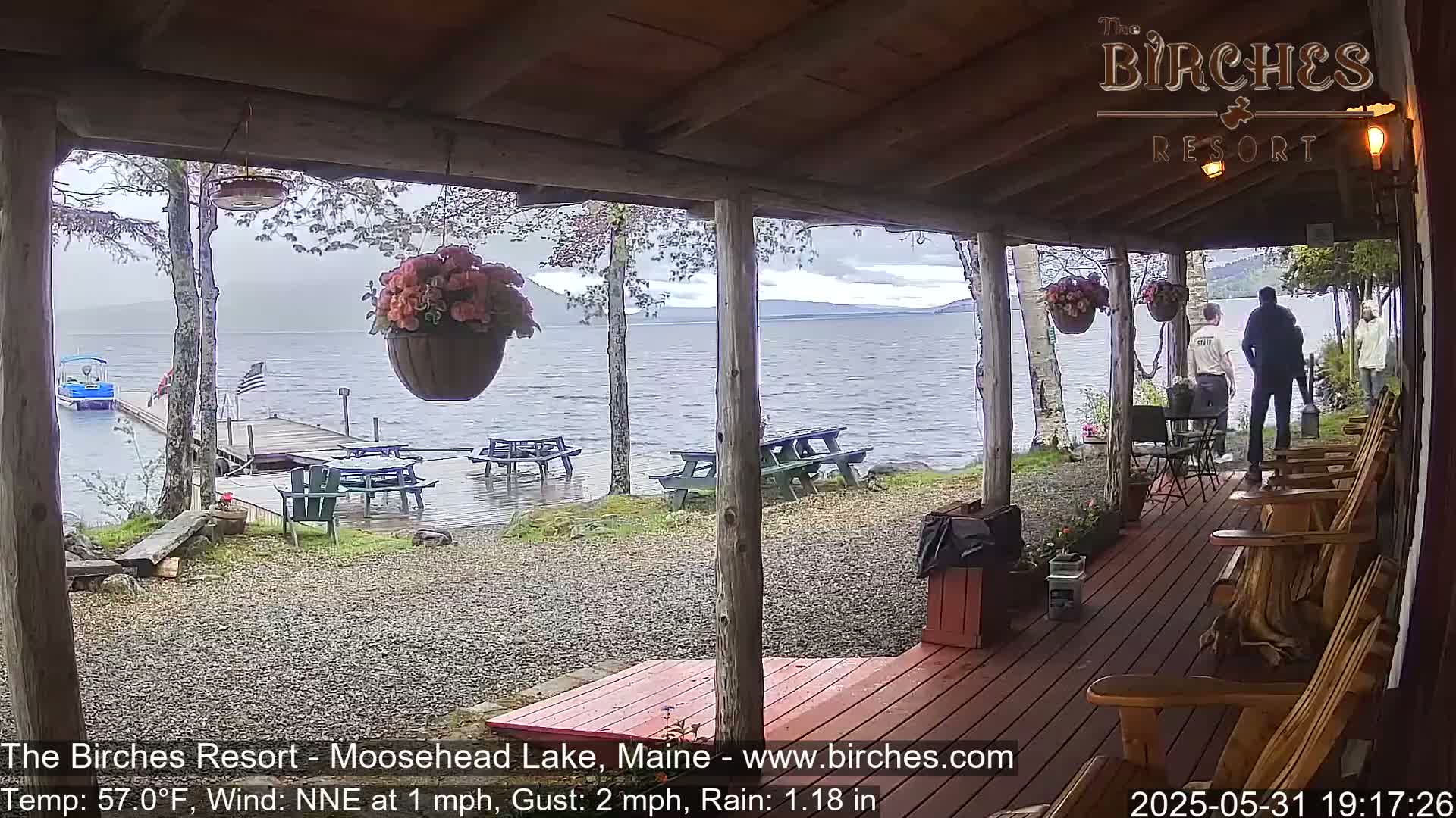 A covered porch overlooks a rainy lake with picnic tables and a dock visible, and several people stand near the water's edge.