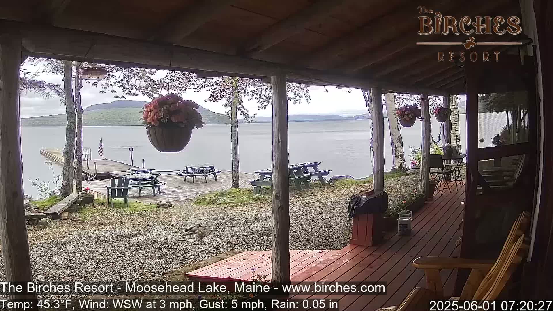 A lakeside porch with picnic tables and hanging flower baskets overlooks a calm lake under a cloudy sky with light rain.