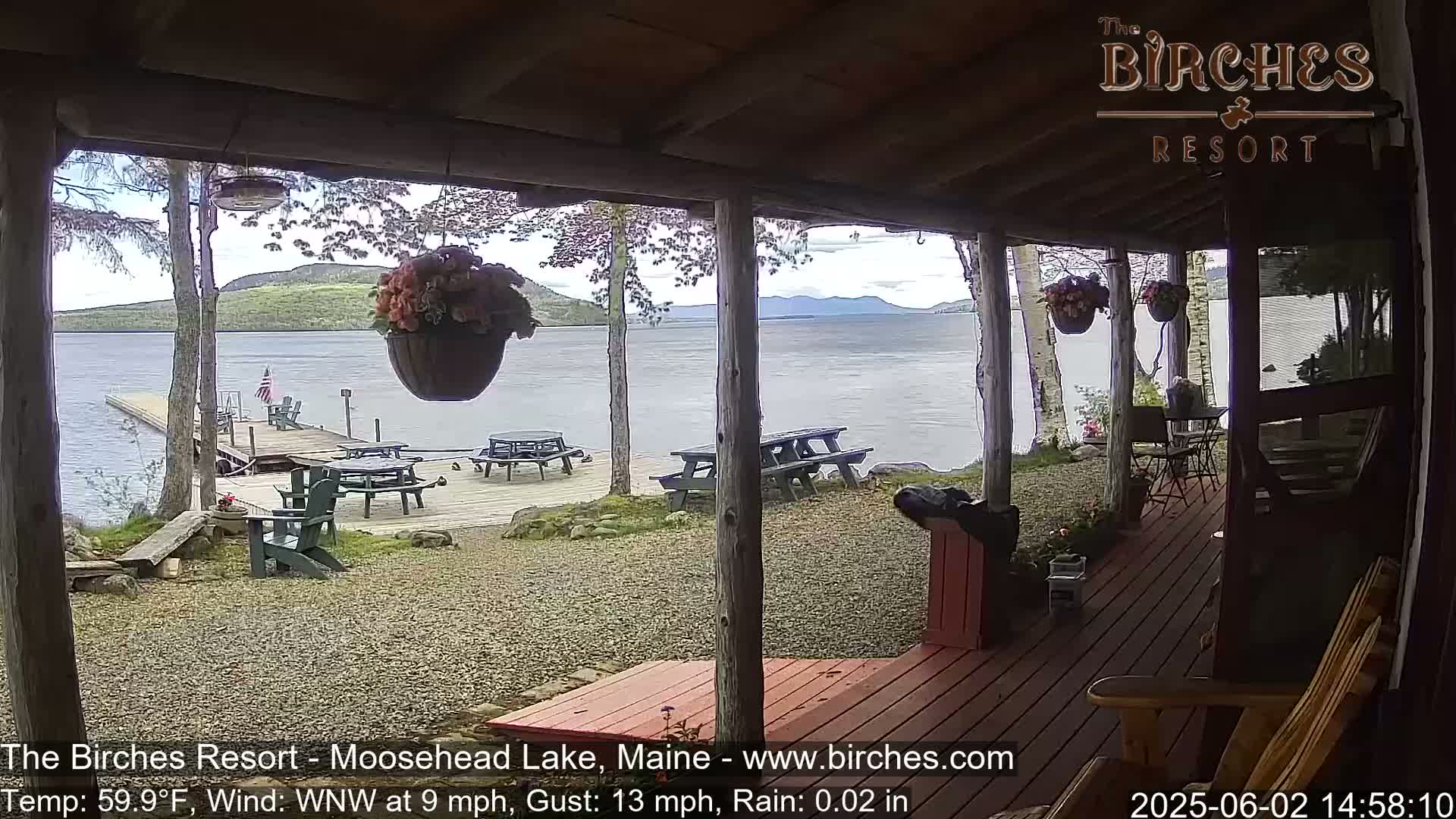 A view from a porch shows a calm lake with mountains in the distance, picnic tables on a dock, and hanging flower baskets, under a slightly rainy sky.