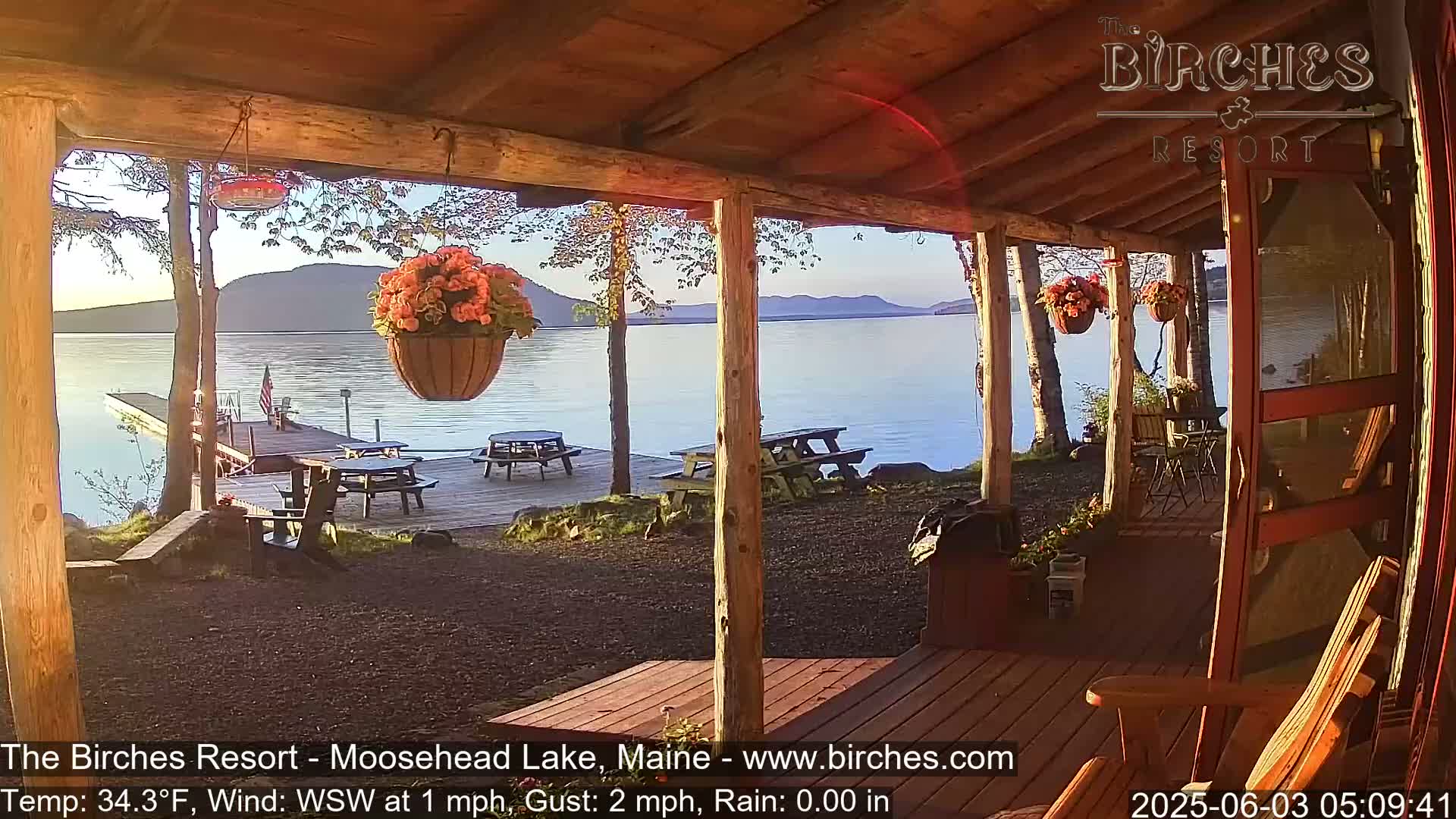 A calm, sunny morning view from a wooden porch overlooking a lake with picnic tables on a dock, and hanging flower baskets.