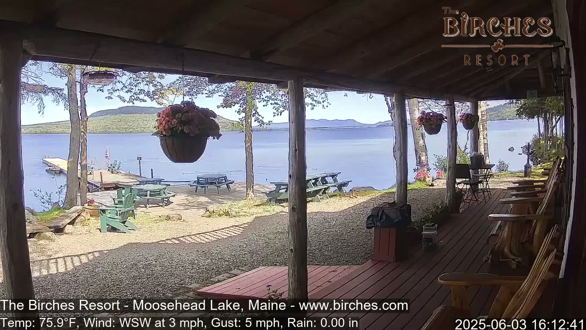 A lakeside porch with wooden chairs and picnic tables overlooks a calm lake under a sunny sky.