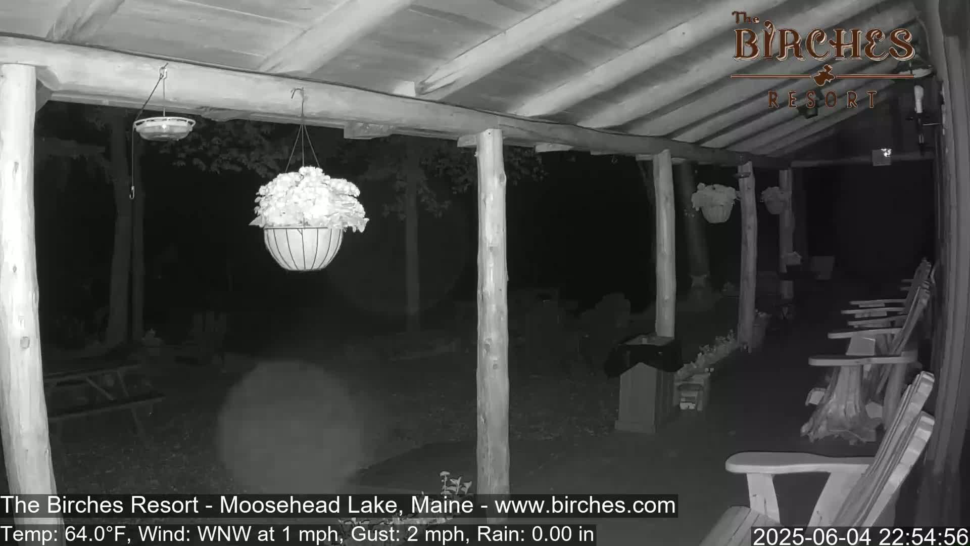 A nighttime, monochrome view of a covered porch with hanging planters, chairs, and a mostly dark yard under a calm, rainless sky.