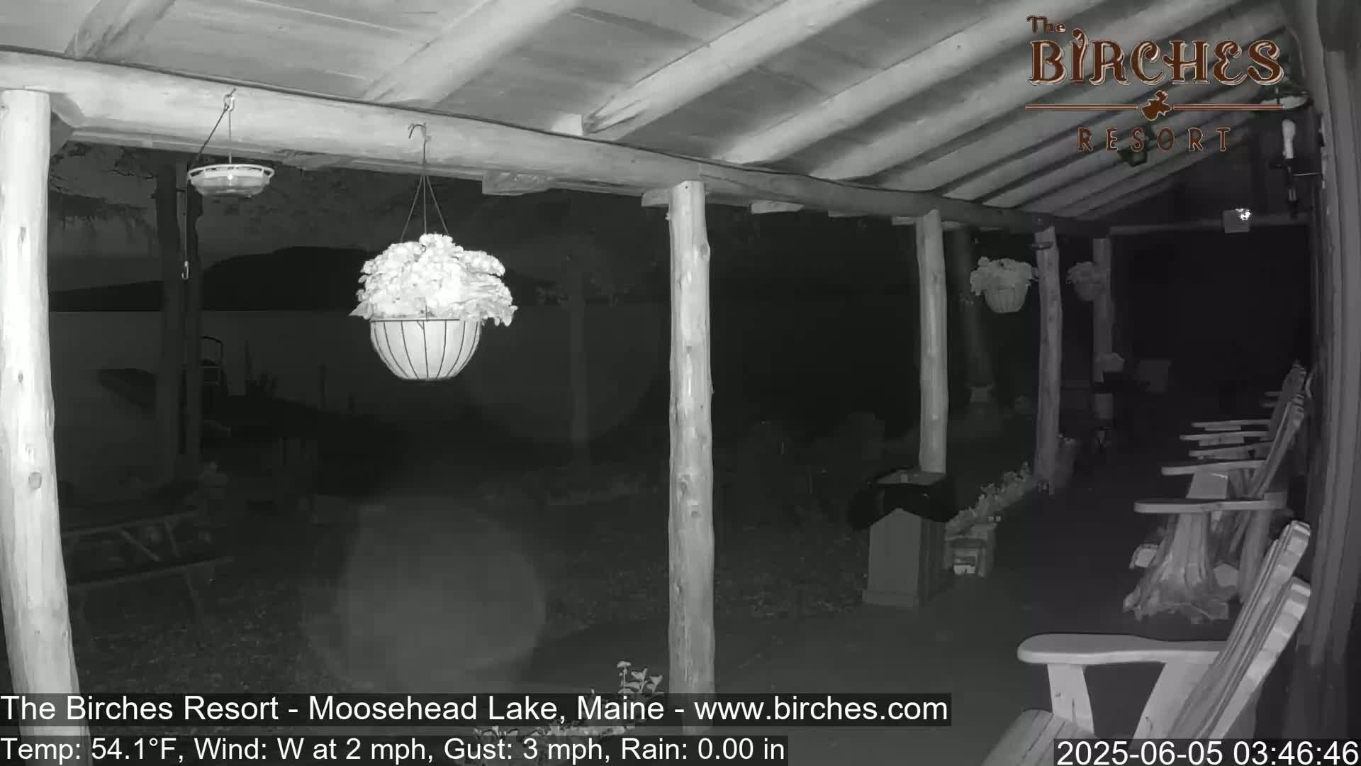 A nighttime, low-light view of a covered porch with hanging plants, wooden posts, and several Adirondack chairs, under conditions of light rain and a westerly wind.