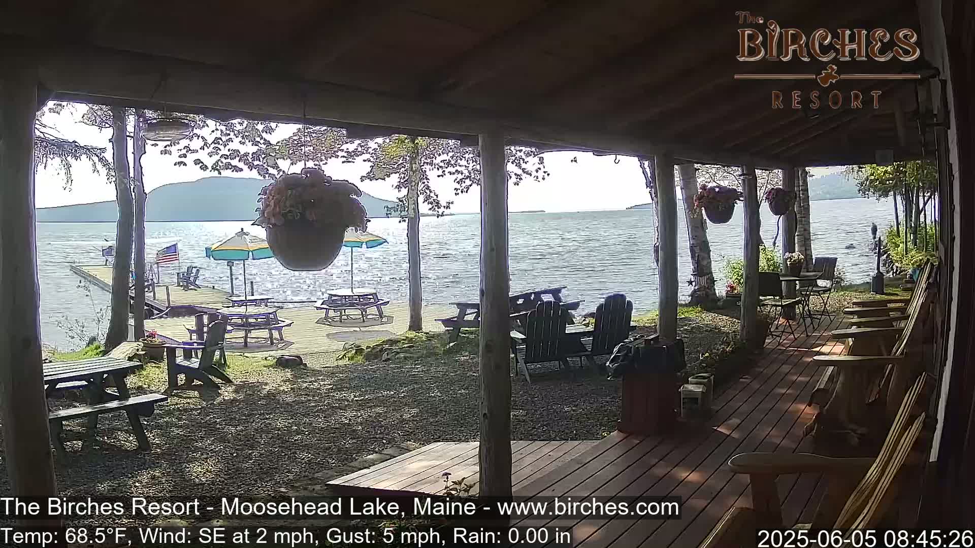 A lakeside porch with picnic tables and Adirondack chairs overlooks calm water under a partly sunny sky.