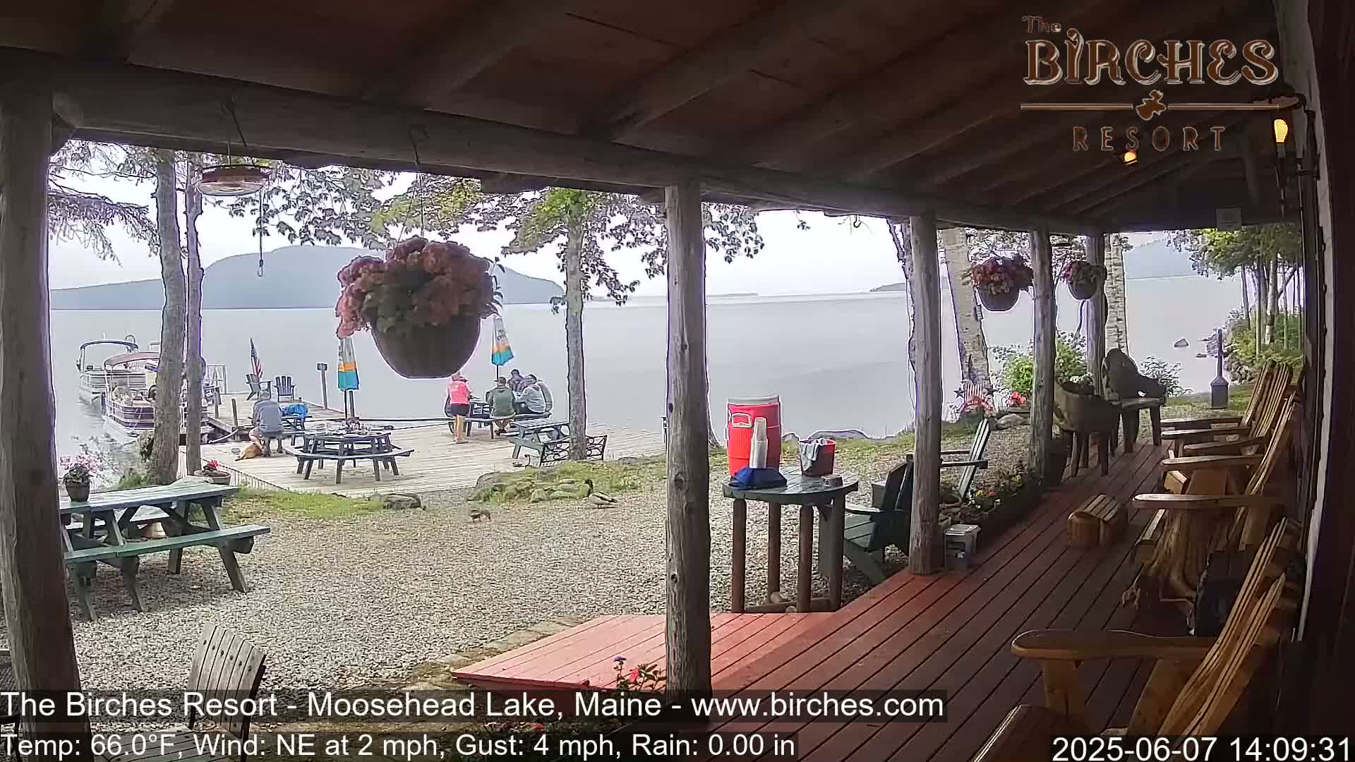 From a porch overlooking a calm lake, several people sit at picnic tables near a dock with boats, under a lightly rainy sky.