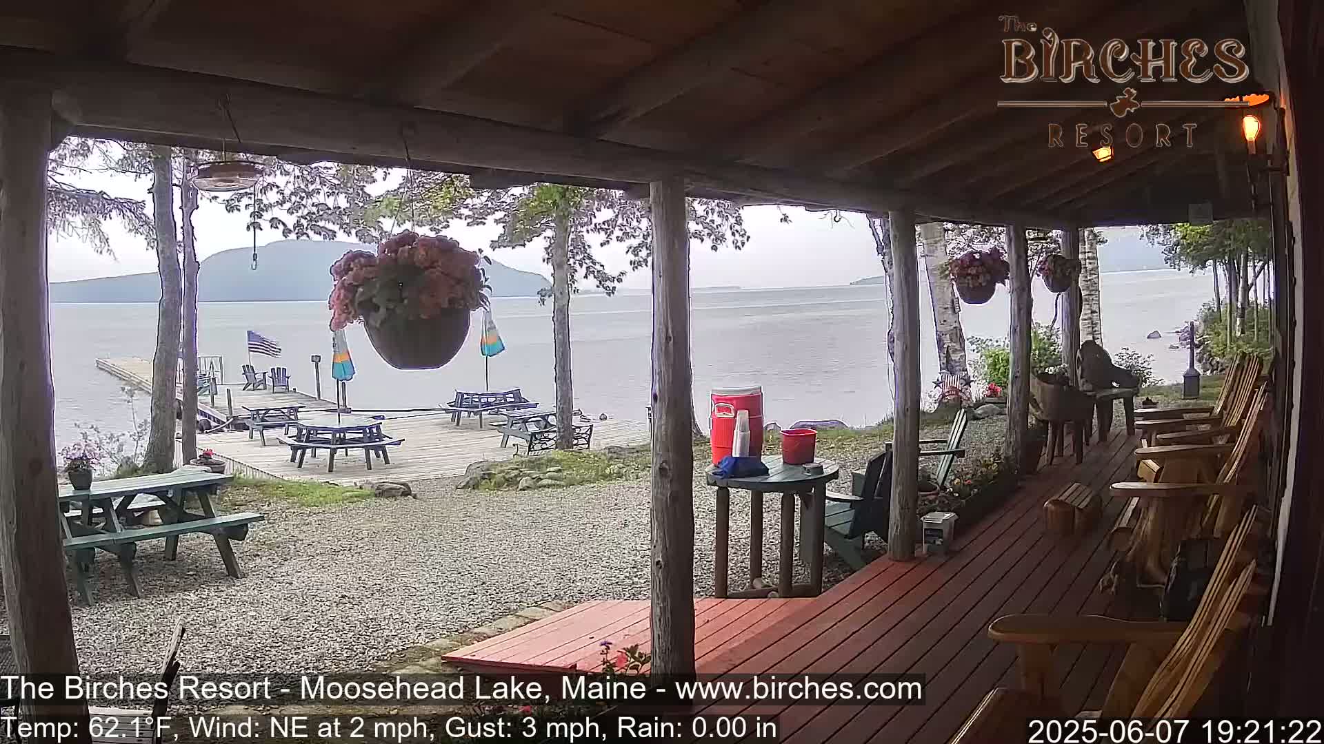 A lakeside porch with picnic tables and Adirondack chairs overlooks a calm lake under a cloudy sky.