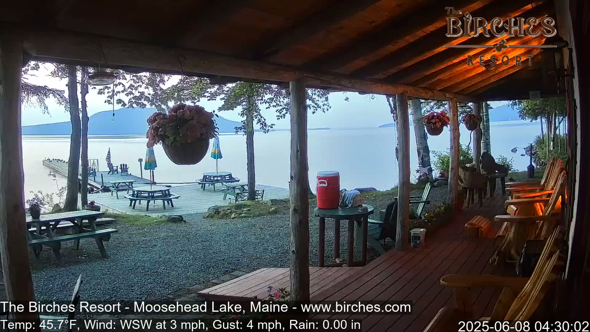 A lakeside porch with wooden furniture overlooks a calm lake and dock under an overcast sky.