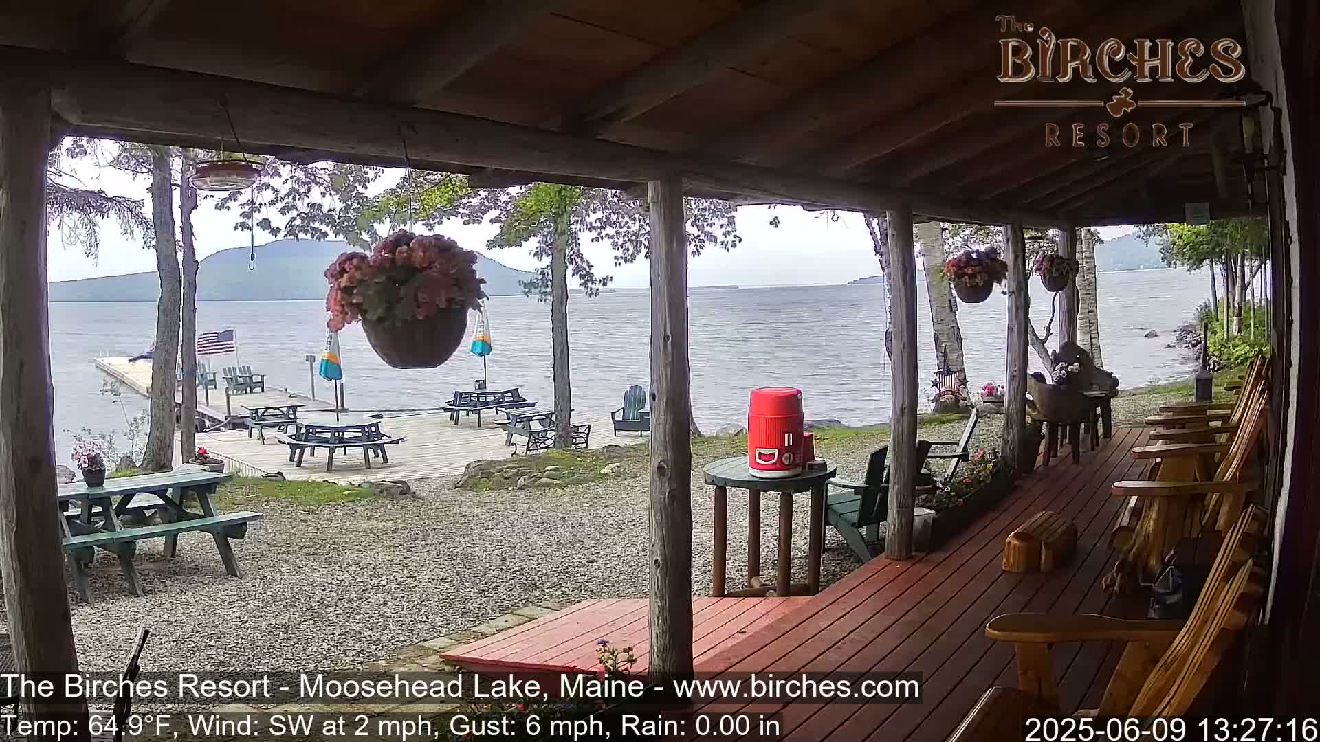 A lakeside porch with wooden chairs and picnic tables overlooks a calm lake under an overcast sky.