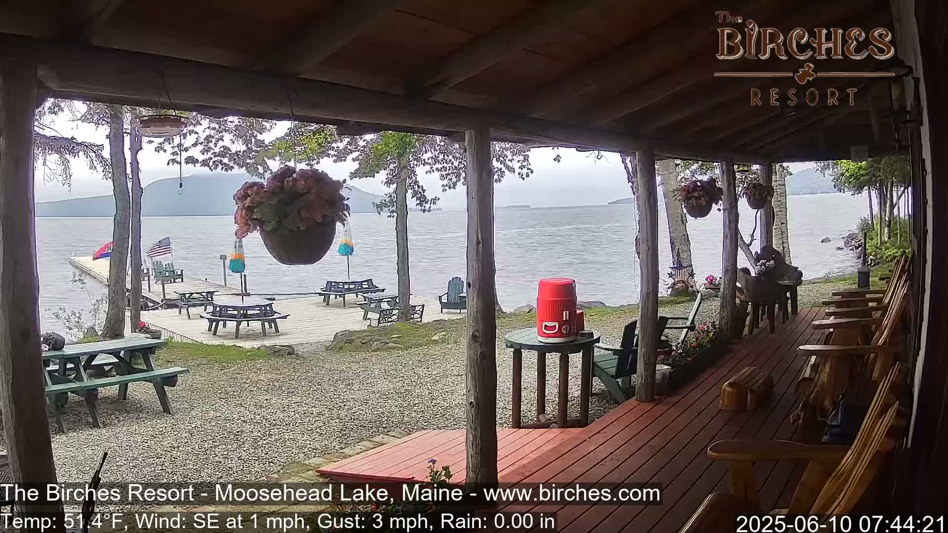 A view from a covered porch shows a lake with mountains in the distance, picnic tables on a gravel area by the water, and wooden chairs on the porch under an overcast sky.