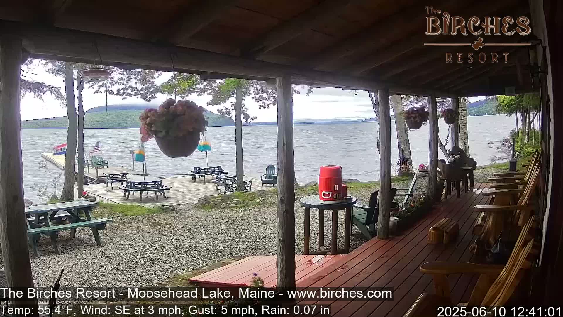 A lakeside porch with wooden chairs and picnic tables overlooks a calm lake under a lightly rainy sky.