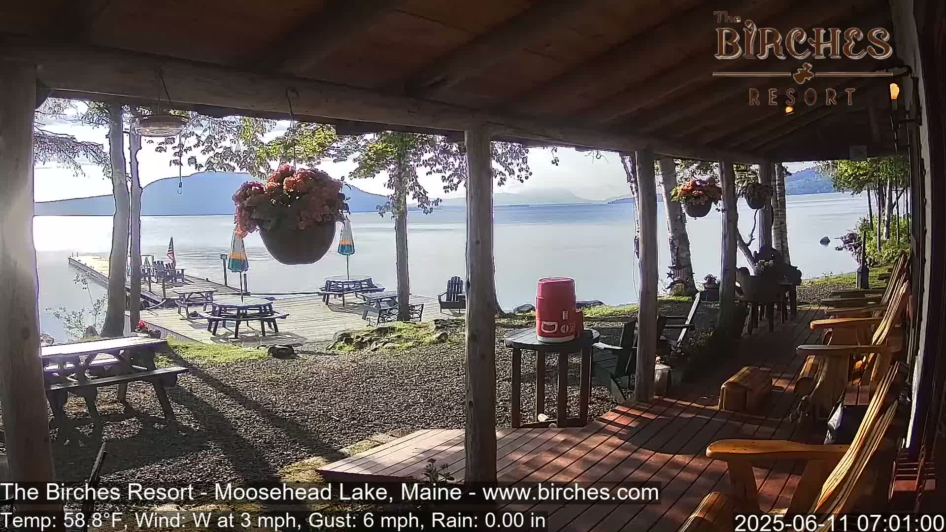 A lakeside porch with wooden chairs and picnic tables overlooks a calm lake under a partly sunny sky, with light rain.