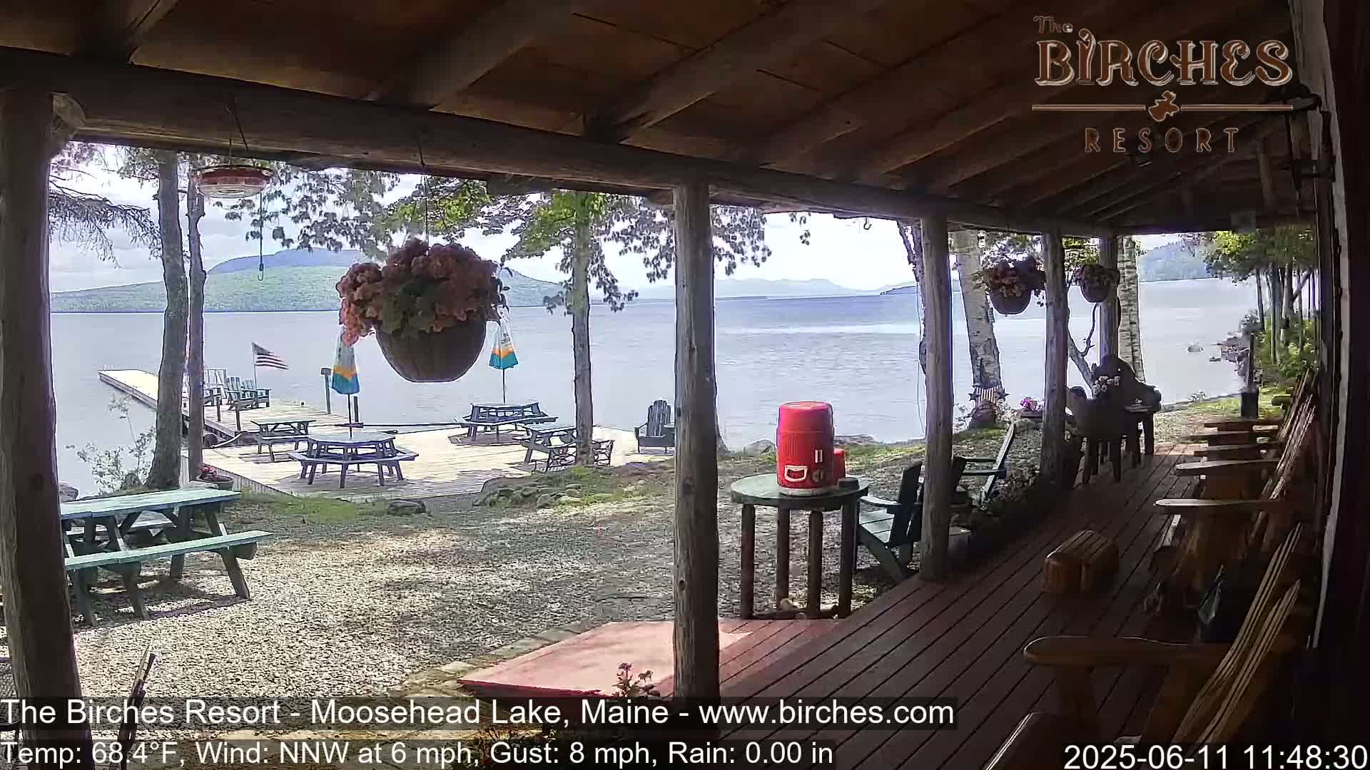A lakeside porch with wooden furniture overlooks a calm lake and a dock with picnic tables, under partly sunny skies.