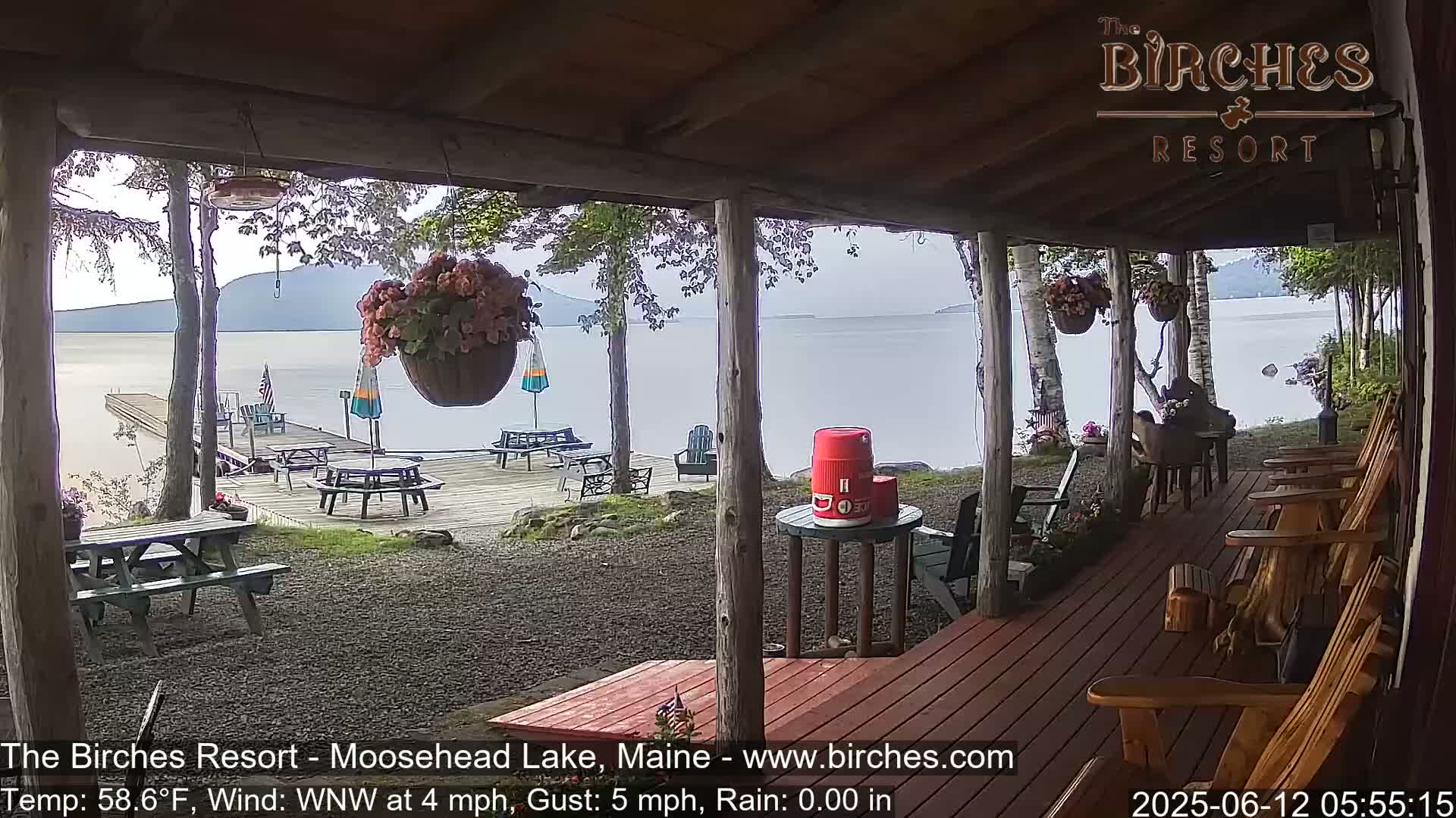 A porch overlooking a calm lake with picnic tables and Adirondack chairs on a deck, under a mostly cloudy sky.