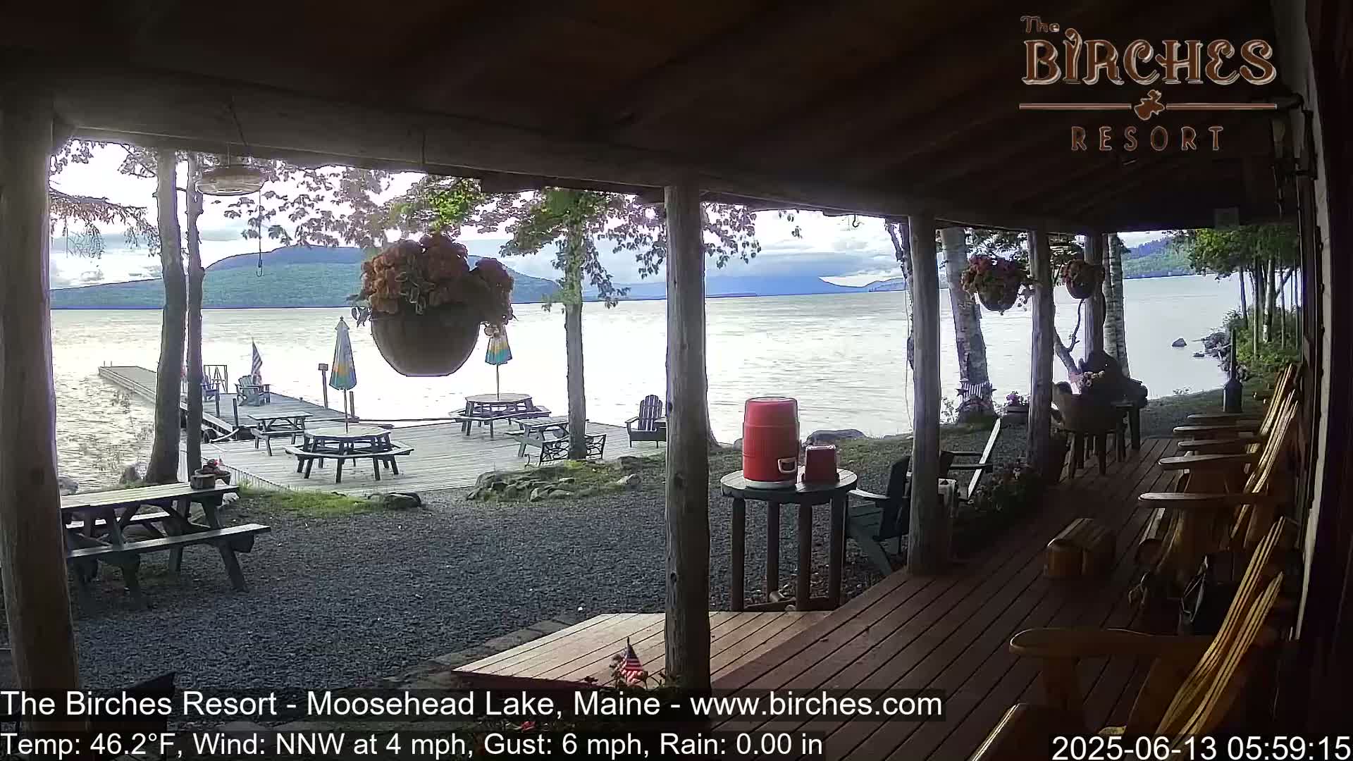 A lakeside porch with wooden chairs and picnic tables overlooks a calm lake and distant mountains under a partly cloudy sky.