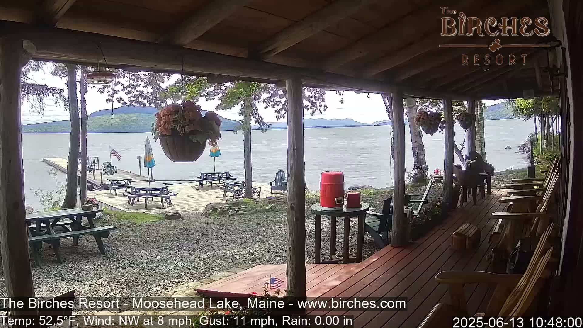 From a covered porch overlooking a calm lake, a wooden deck with picnic tables is visible, with hanging flower baskets and Adirondack chairs.
