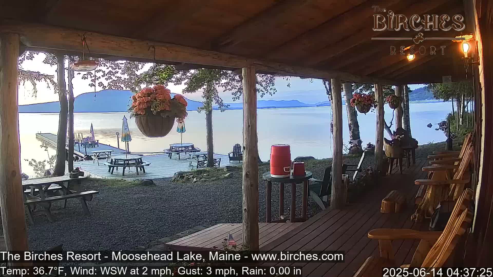 A calm lake view is seen from a wooden porch with picnic tables and rocking chairs, under a partly cloudy sky.