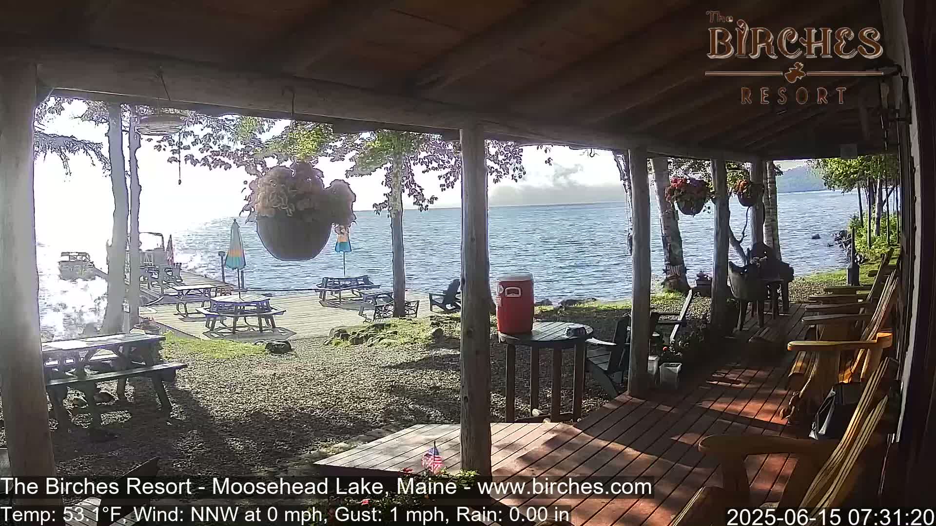 A view from a porch overlooking a calm lake with picnic tables on a gravel area and several wooden rocking chairs on the porch itself, under a partly sunny sky.