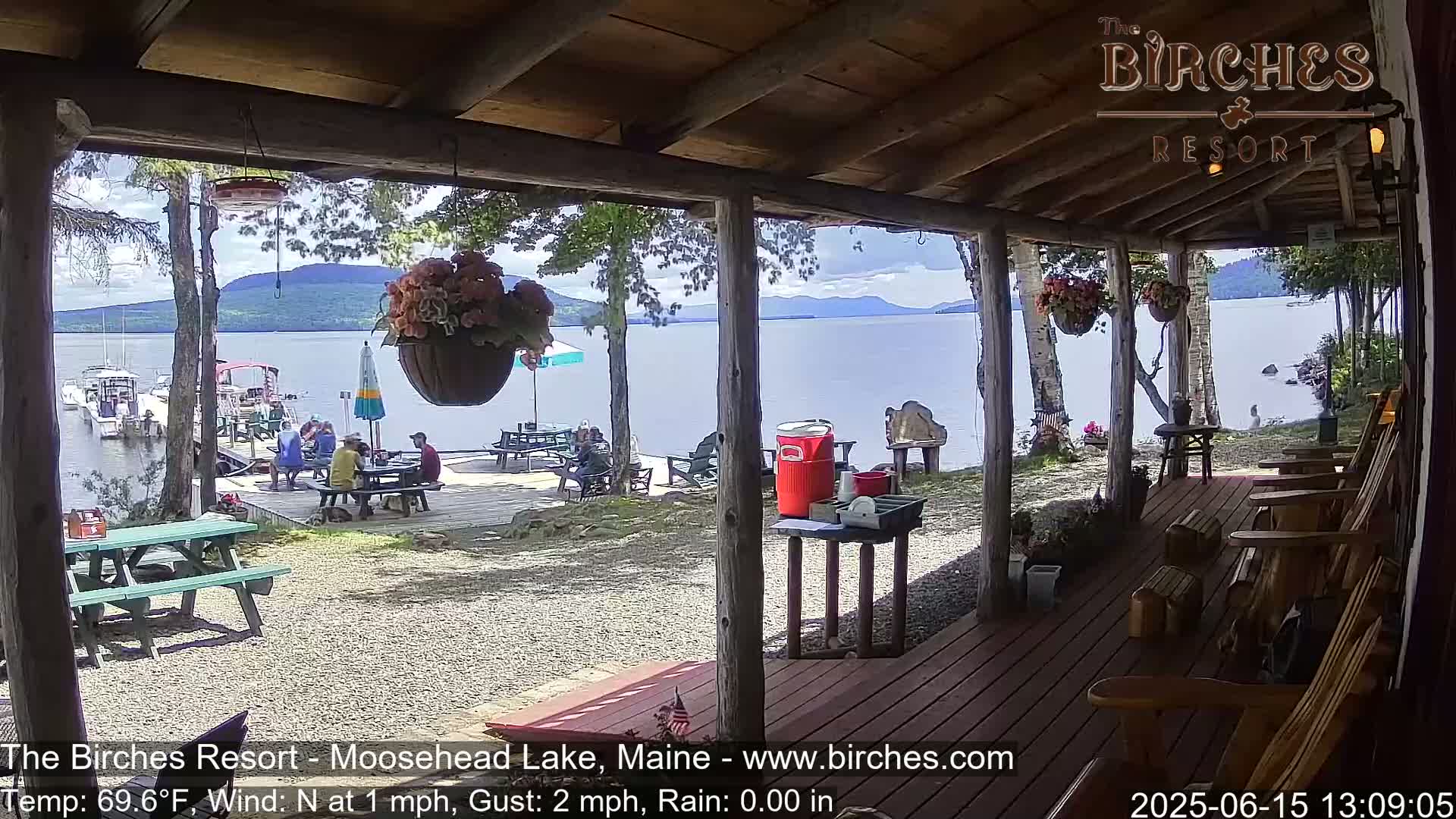 A lakeside porch with rocking chairs overlooks a calm lake and a small beach with people sitting at picnic tables, under partly cloudy skies.