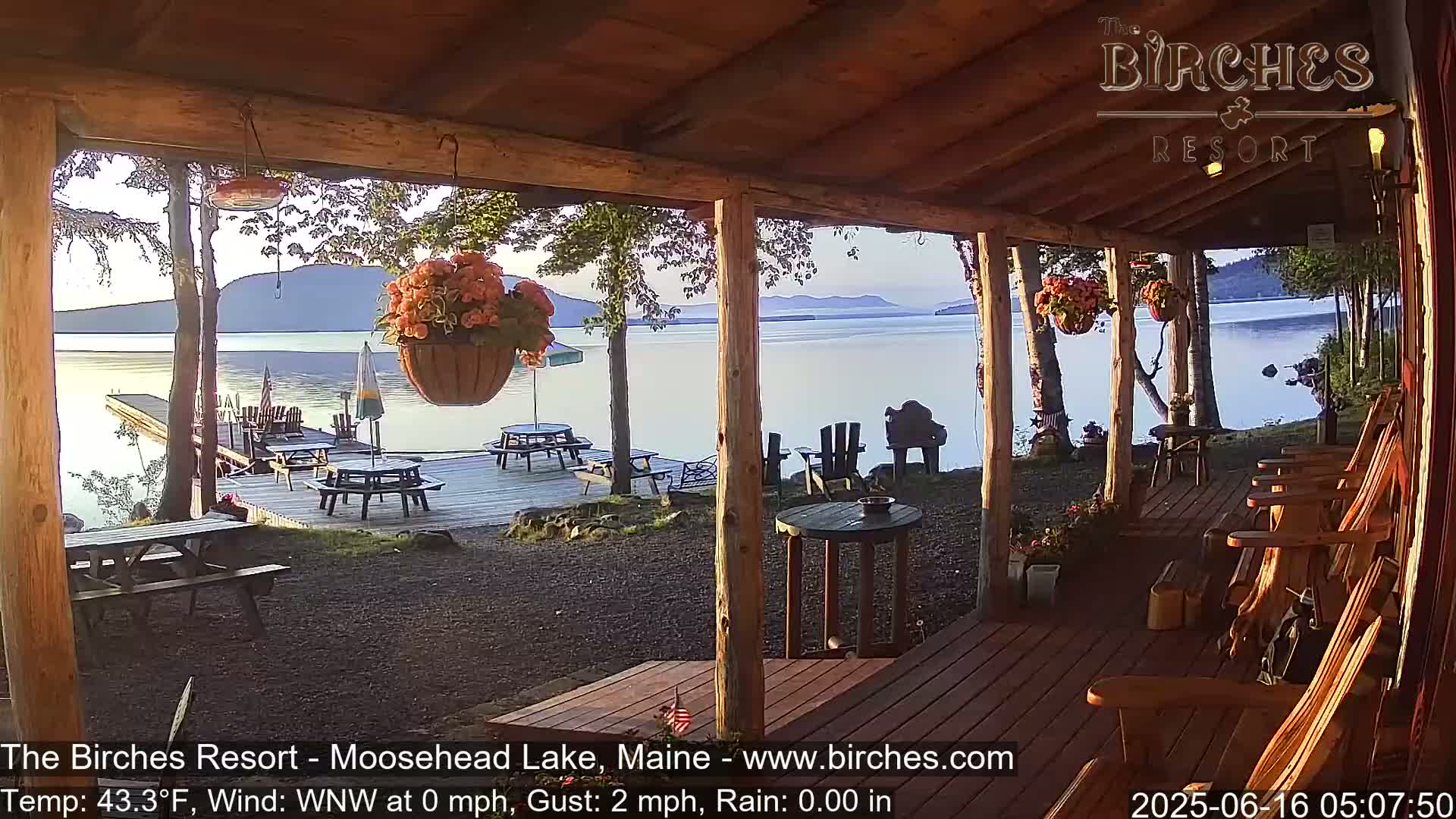 A calm lake view is seen from a wooden porch with picnic tables and rocking chairs, under a clear morning sky.