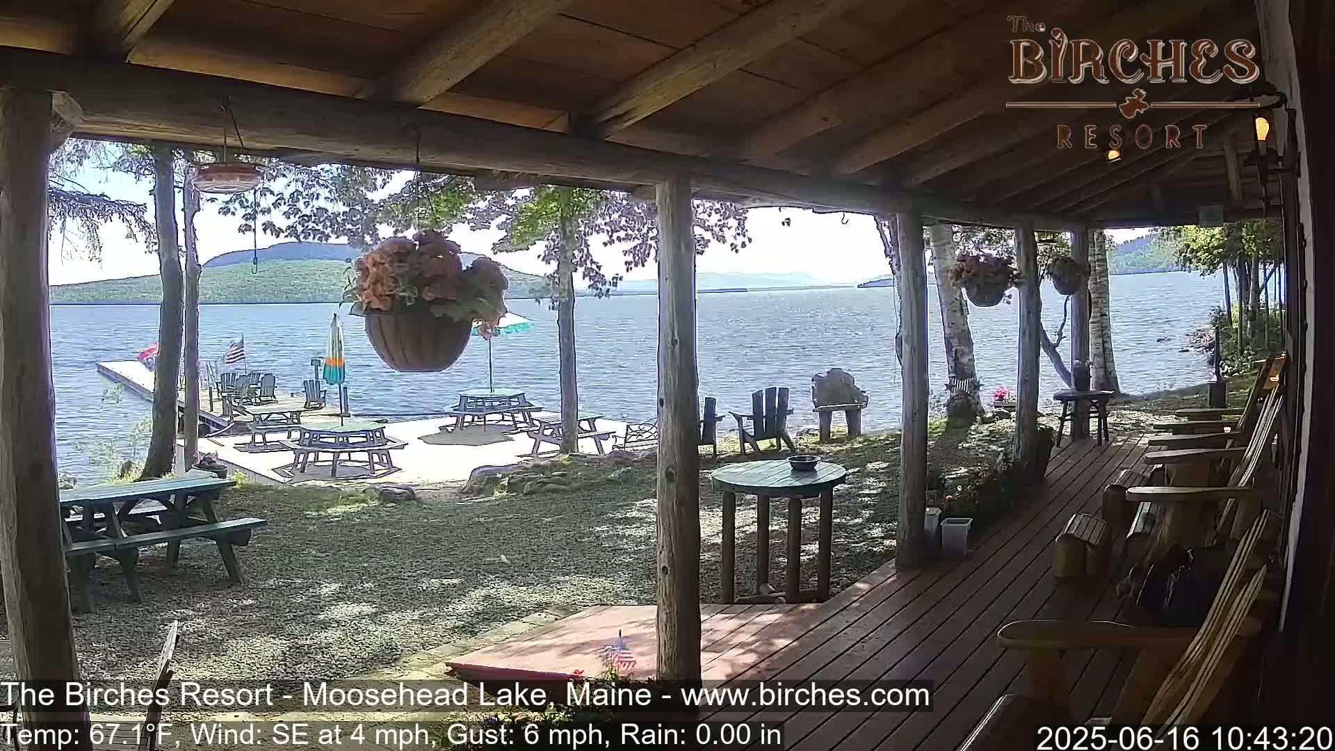 A lakeside porch with picnic tables and Adirondack chairs overlooks a calm lake under a partly sunny sky.