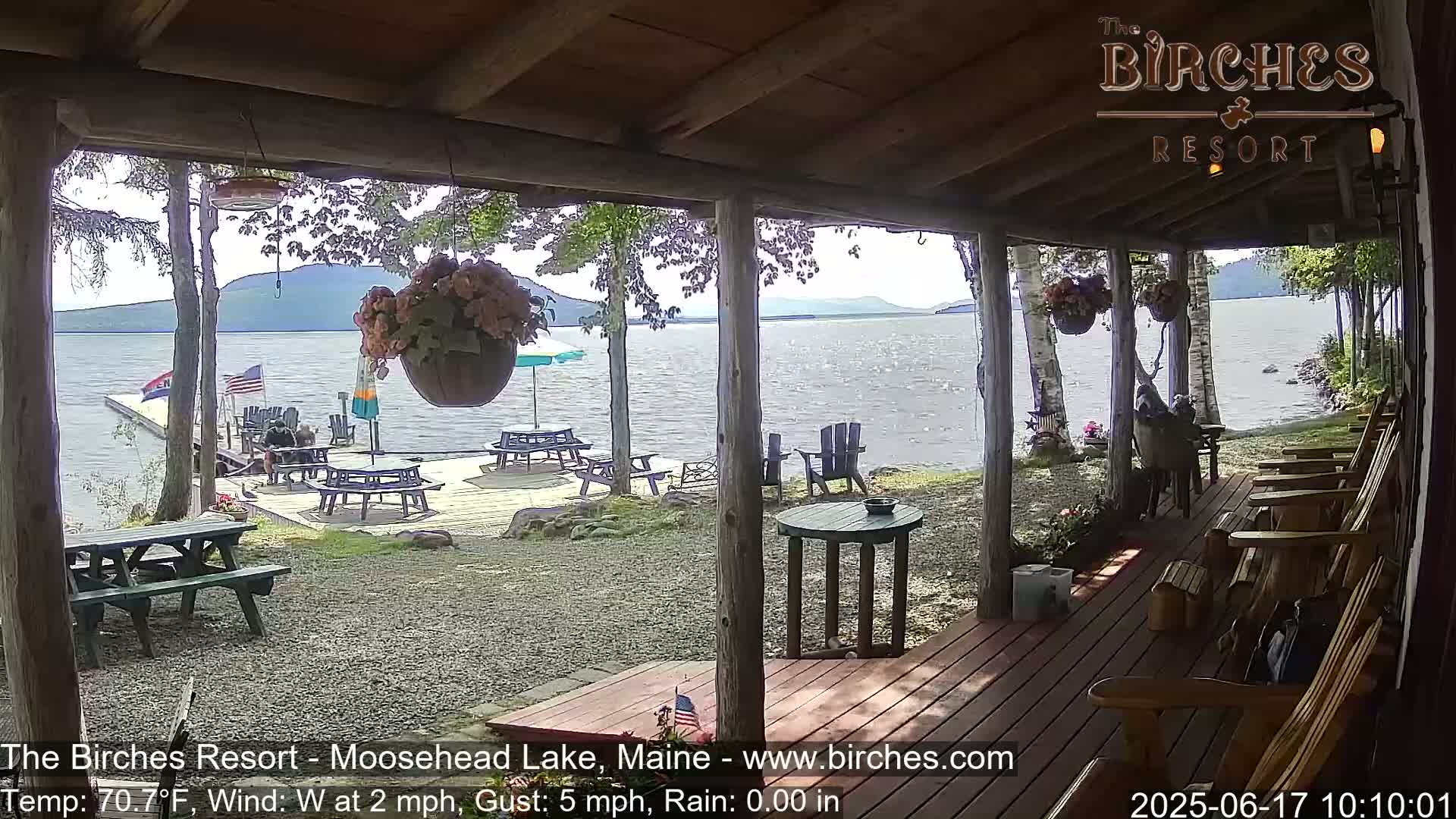 A lakeside porch with picnic tables and Adirondack chairs overlooks a calm lake under a partly sunny sky.