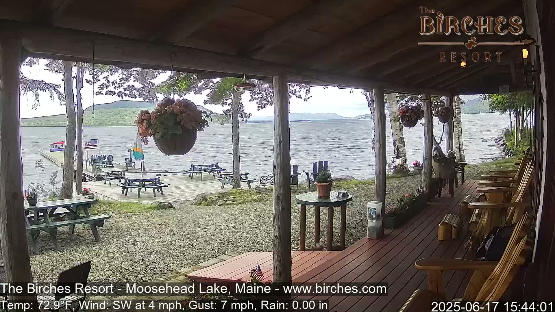 A covered porch overlooks a lake with picnic tables and chairs on a deck below, under partly cloudy skies with a gentle southwest wind.