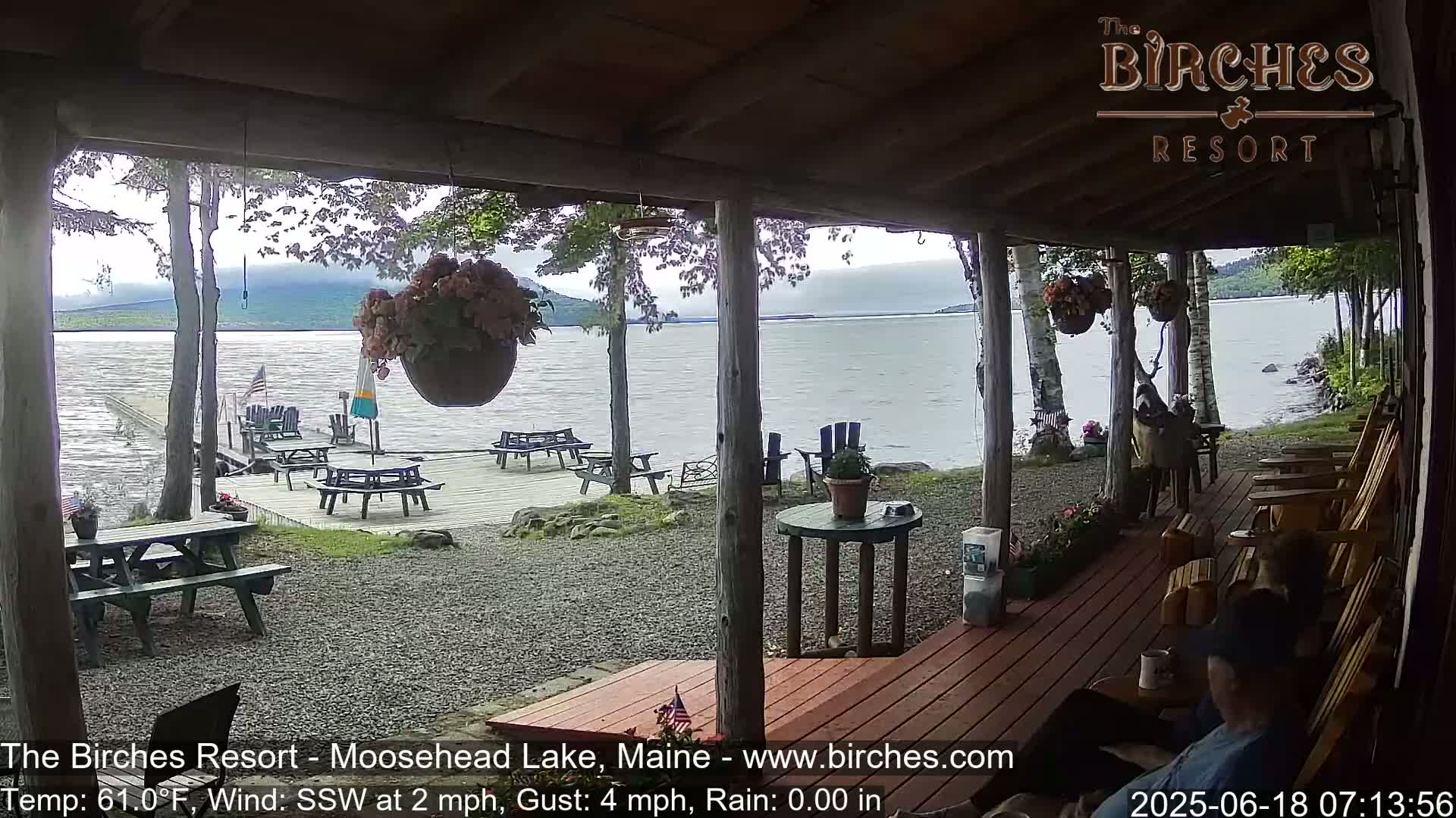 A covered porch overlooks a calm lake with picnic tables and Adirondack chairs on a gravel shoreline under an overcast sky.