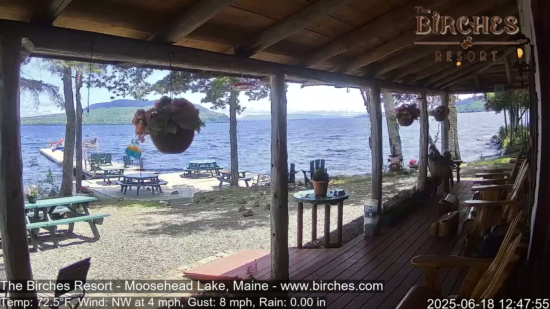 A porch overlooking a lake with a gently waving surface, under a sunny sky, shows picnic tables and chairs on a gravel area leading to a dock, with Adirondack chairs on the porch.