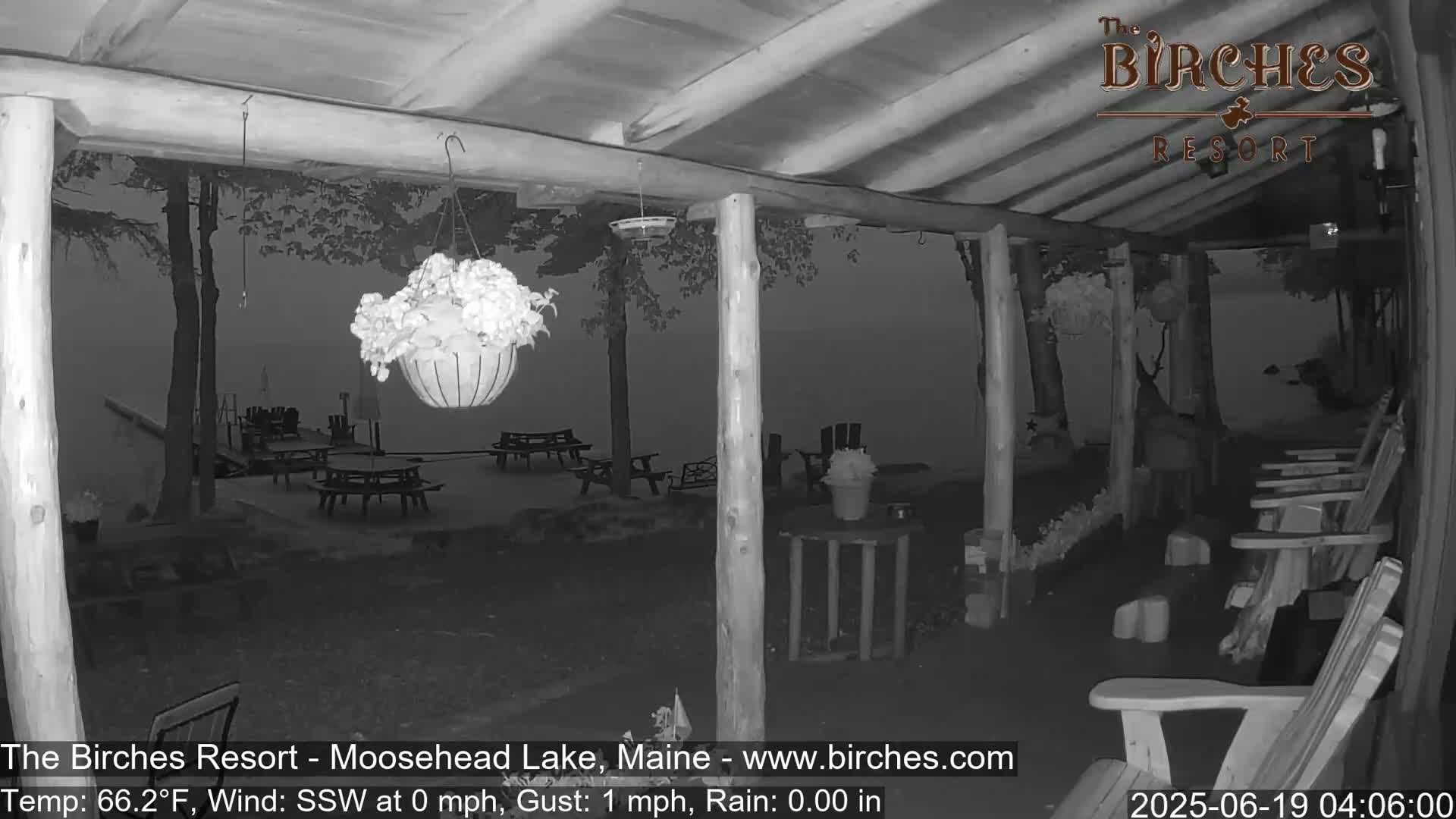 A nighttime view from a covered porch shows a calm lake, picnic tables, and Adirondack chairs in the rain.