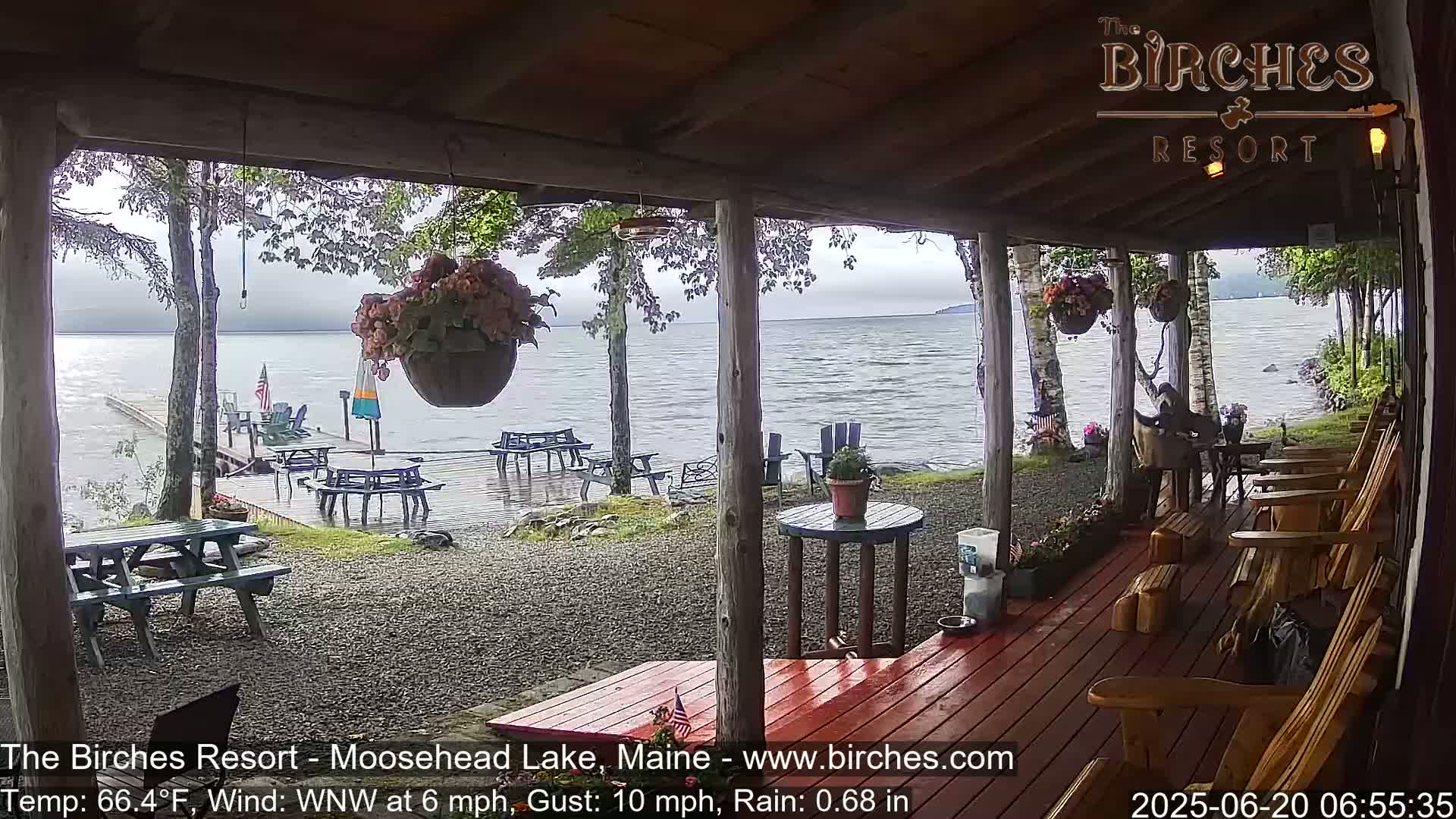 A lakeside porch with wooden chairs and picnic tables, overlooking a rain-swept lake under a cloudy sky.