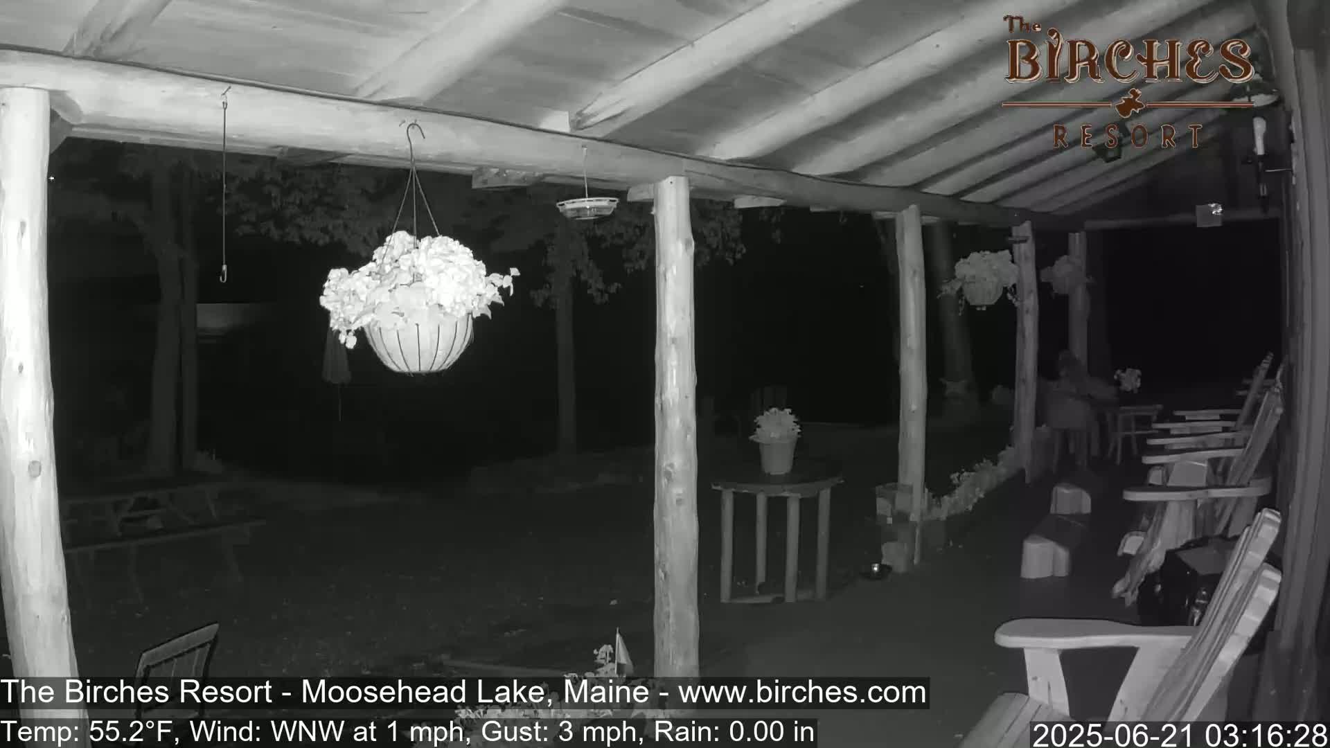 A nighttime, low-light view of a covered porch with hanging plants, a table, and several wooden chairs, under calm, dry conditions.