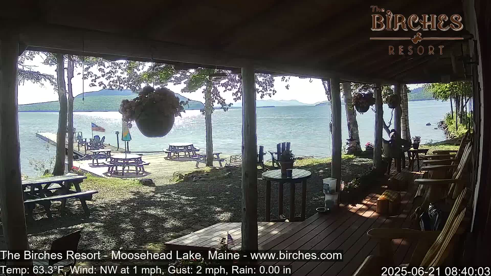 A covered porch overlooks a calm lake with picnic tables and chairs on a deck, under a sunny sky.