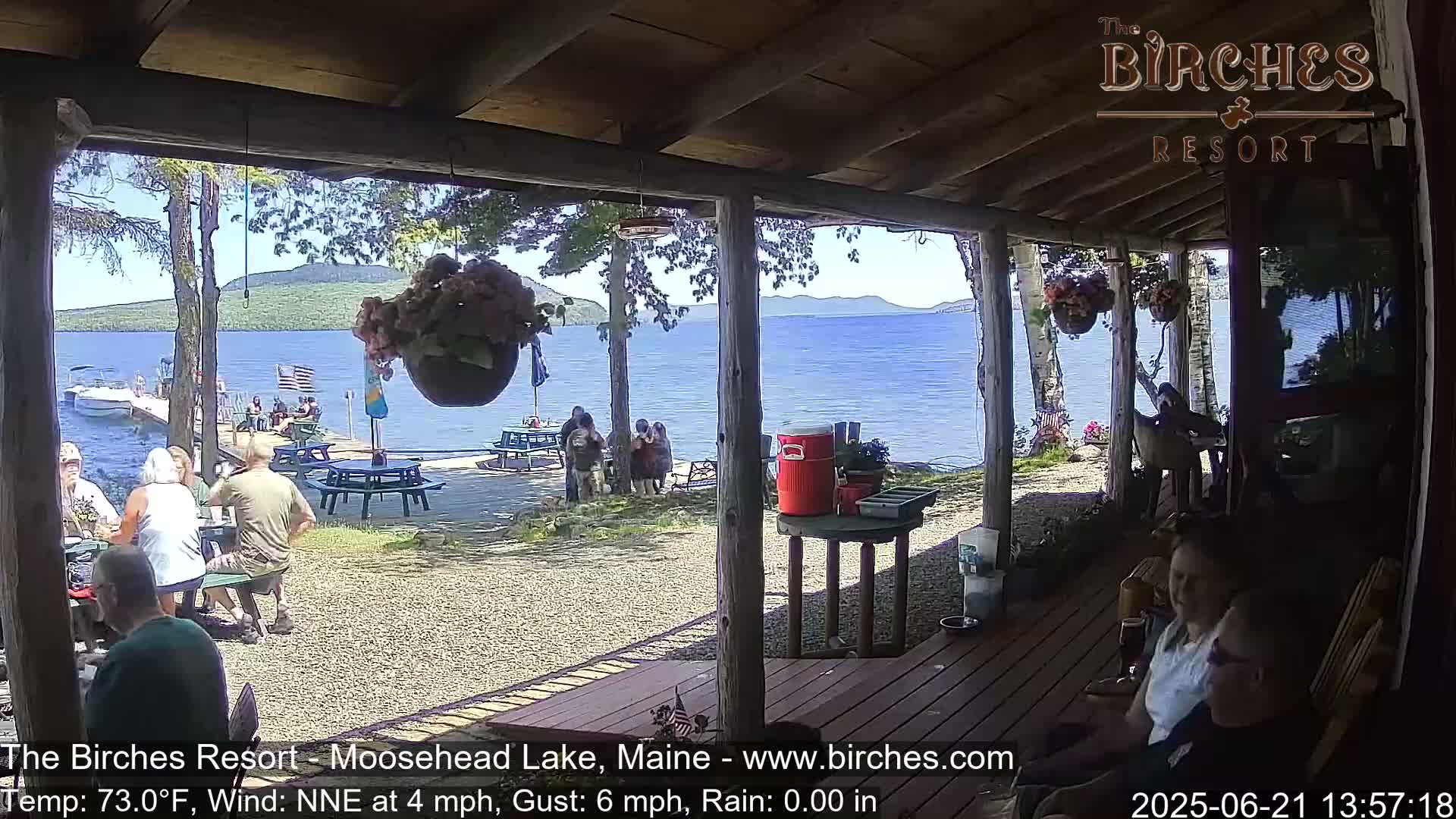 A lakeside porch with people sitting at tables and on chairs, overlooking a calm lake on a sunny day with a light wind.