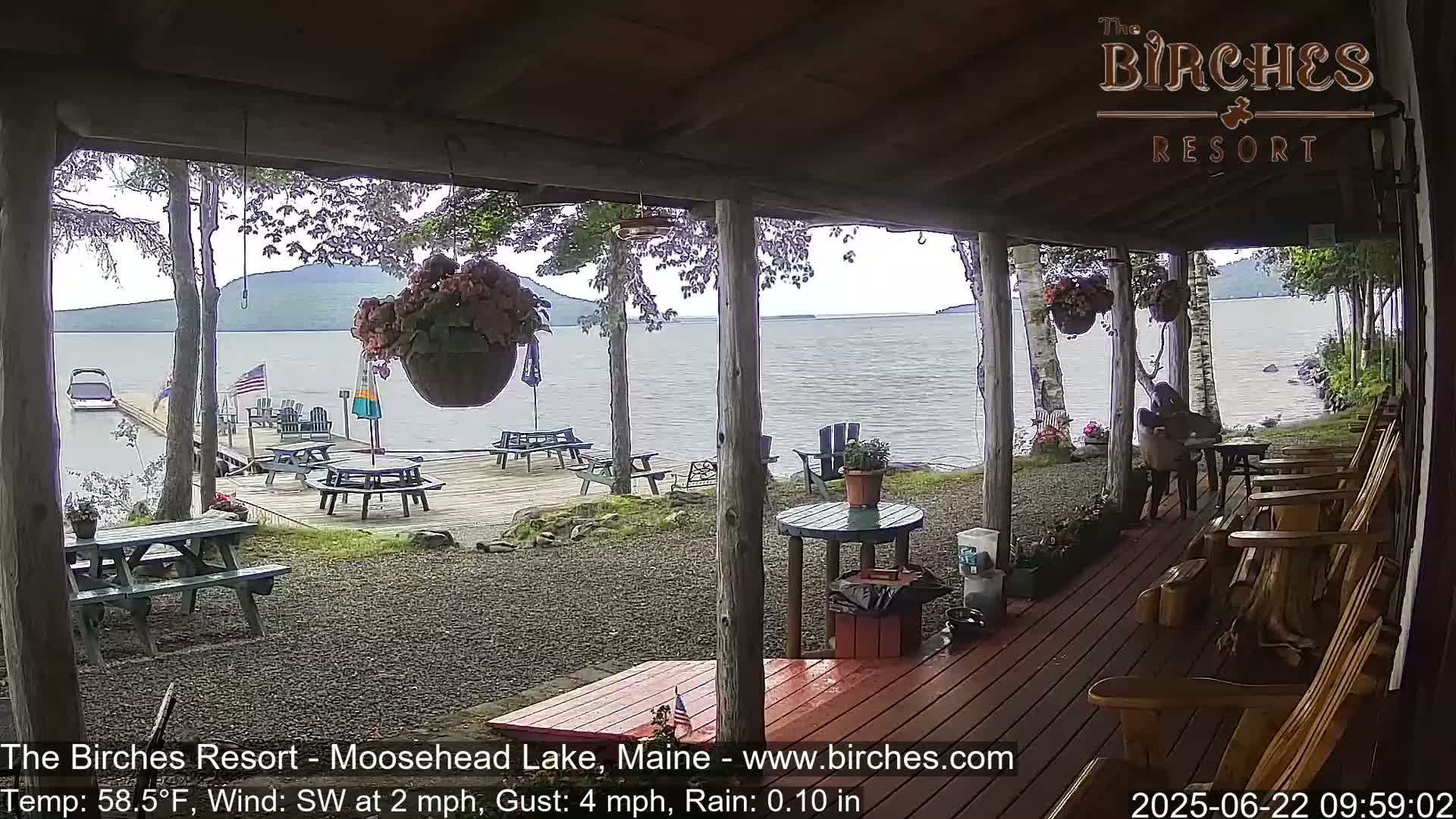 A covered porch overlooks a lake with picnic tables and chairs on a deck, under rainy skies.
