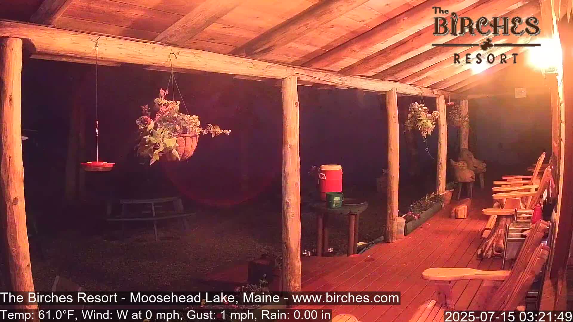 A dimly lit wooden porch at night with hanging flower baskets, rocking chairs, and a bird feeder, under calm conditions.