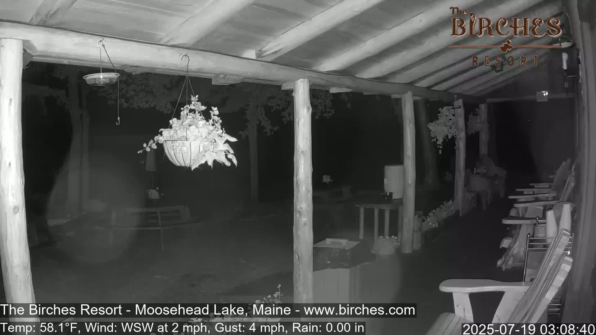 A nighttime, low-light image shows a covered porch with hanging plants, outdoor furniture, and calm weather.
