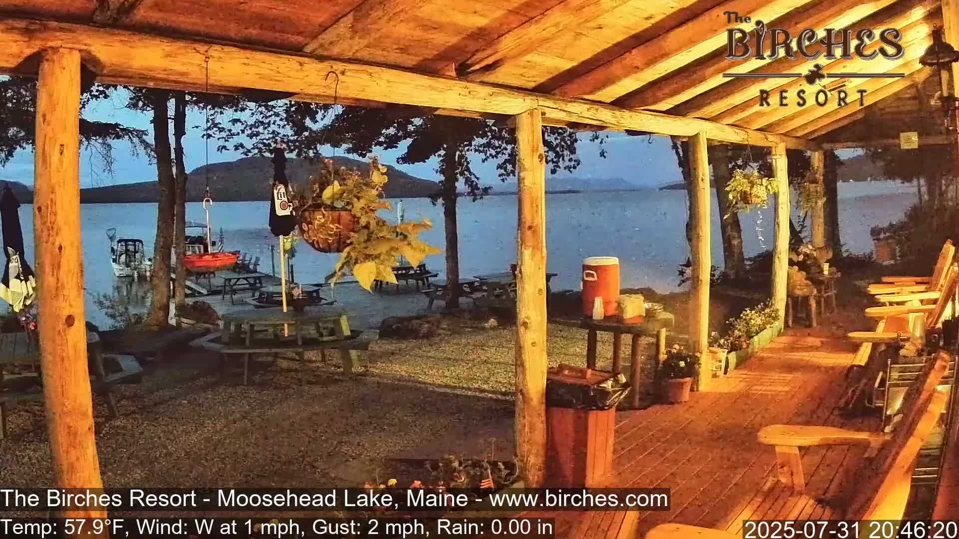 A wooden porch overlooks a calm lake at dusk, with several empty rocking chairs and picnic tables visible in the foreground and background.