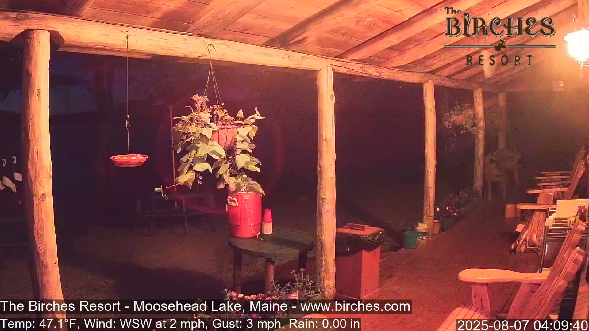 A nighttime view of a wooden porch with hanging plants, a bird feeder, and rocking chairs, under a partly cloudy sky.