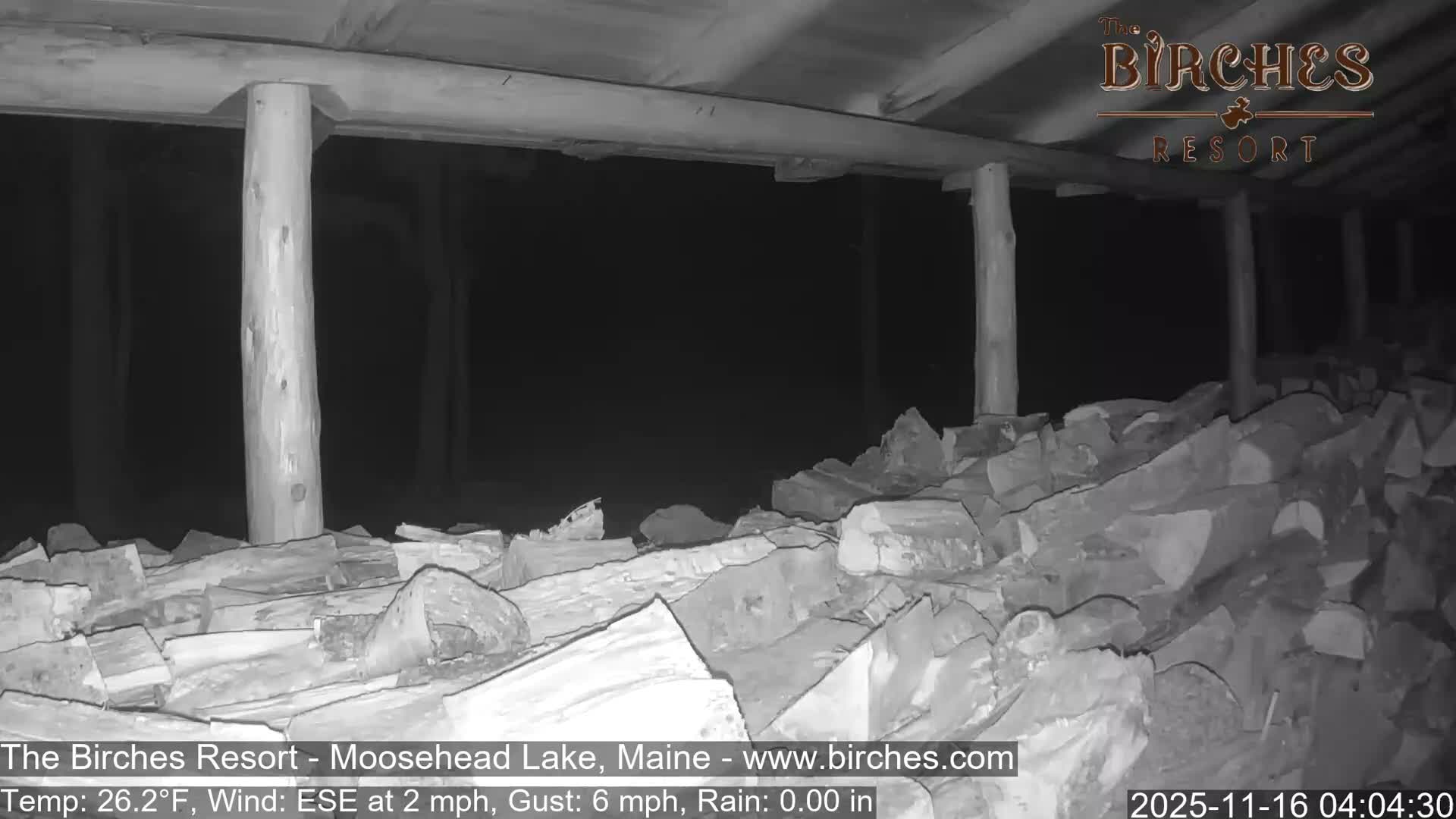 A large pile of split firewood is neatly stacked under a rustic wooden porch or shelter on a clear, dark night.