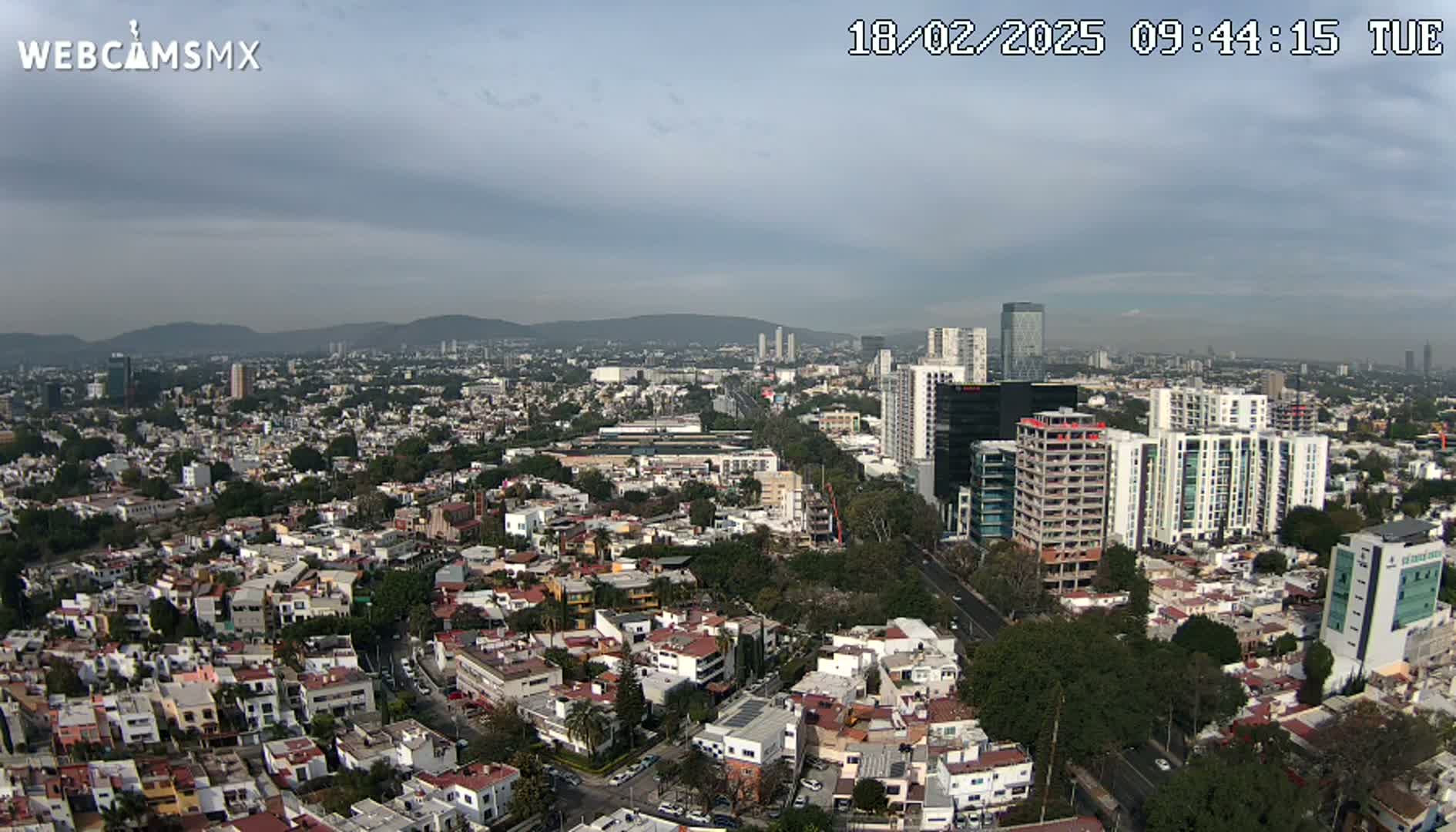 An aerial view shows a sprawling city under a partly cloudy sky, with mountains visible in the distance.