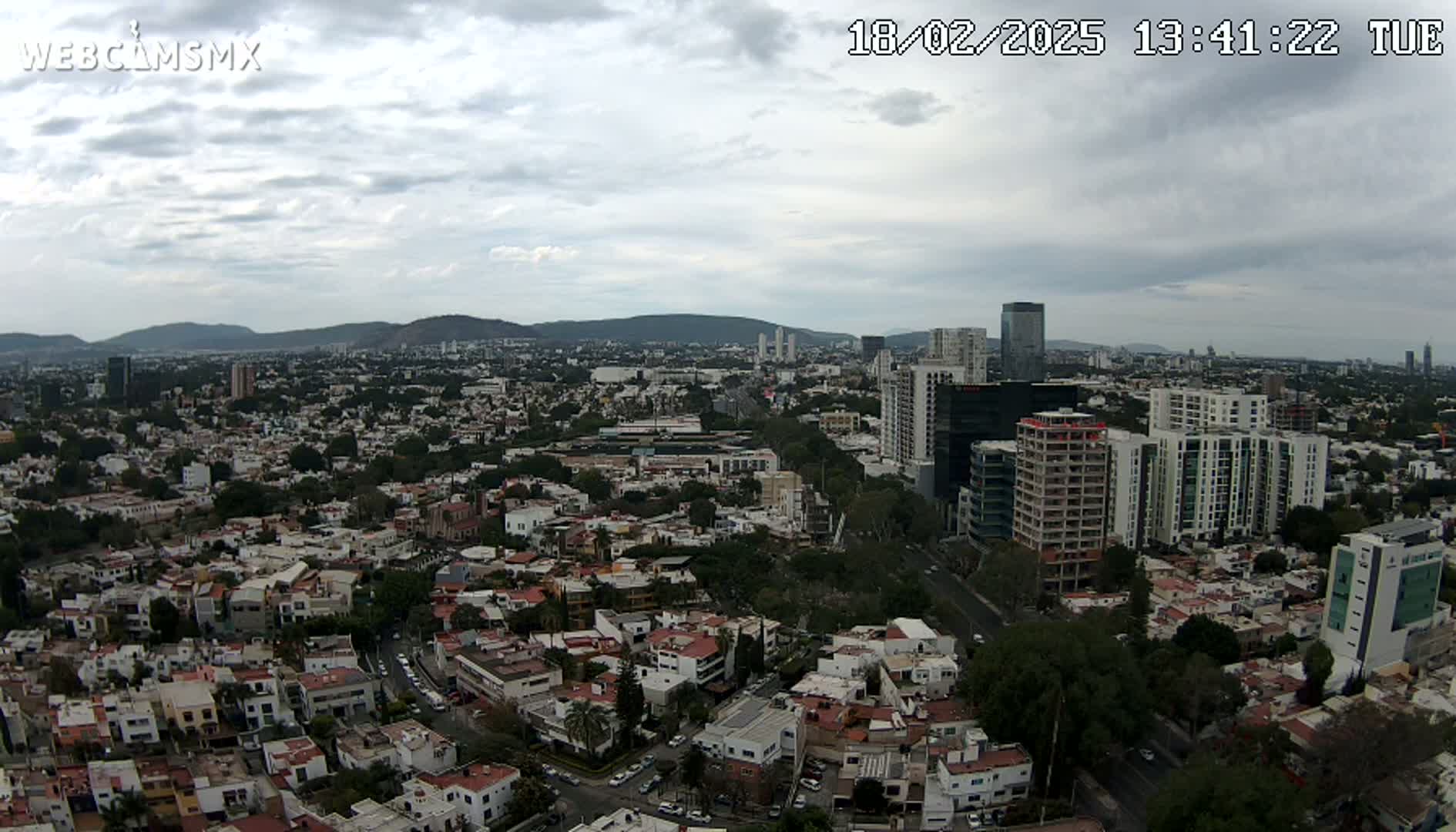An aerial view of a sprawling city under a cloudy sky shows a mix of residential buildings and taller structures, with mountains visible in the distance.