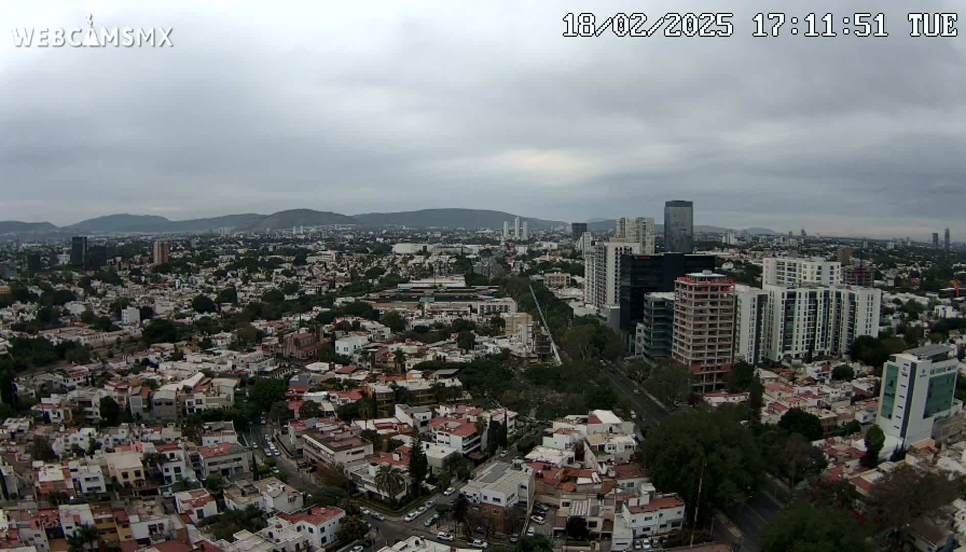 An aerial view of a sprawling city under a cloudy sky shows a mix of low-rise residential buildings and taller structures, with mountains visible in the distance.