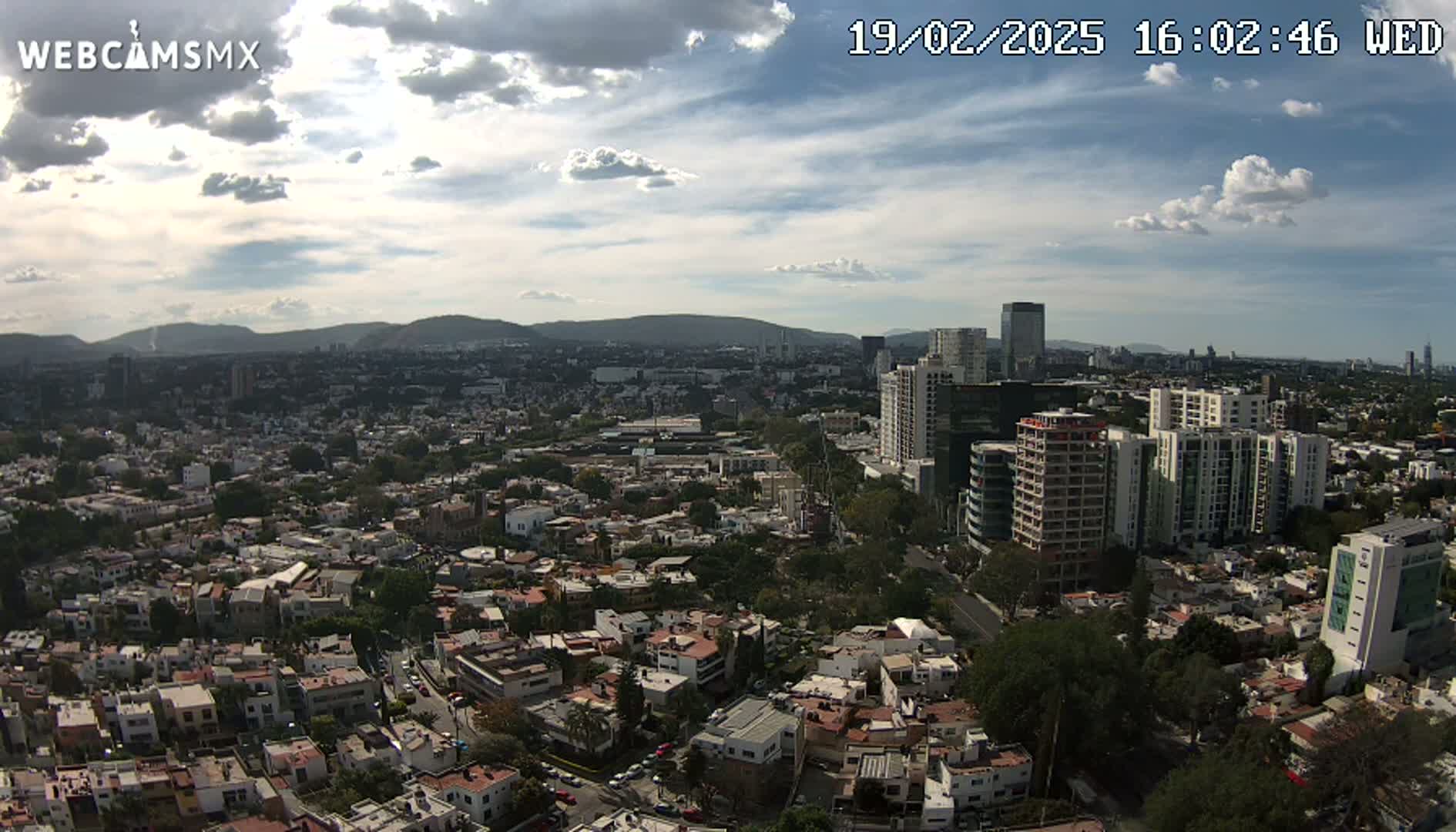 A partly cloudy daytime aerial view shows a sprawling city with numerous residential buildings and taller apartment and office buildings, nestled against a backdrop of distant mountains.