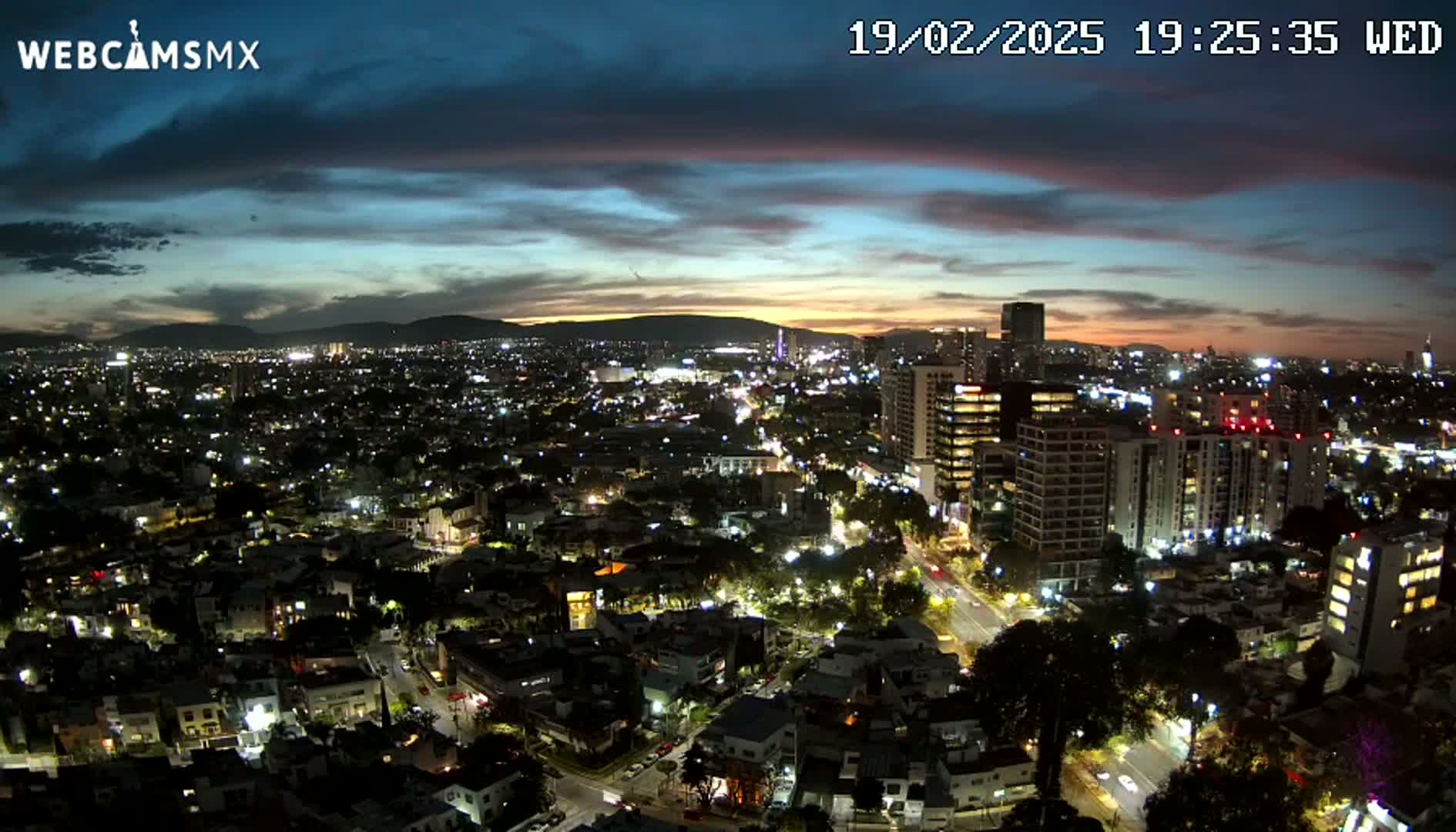 A nighttime aerial view of a city illuminated by streetlights and building lights under a partly cloudy sky with the remnants of a sunset.
