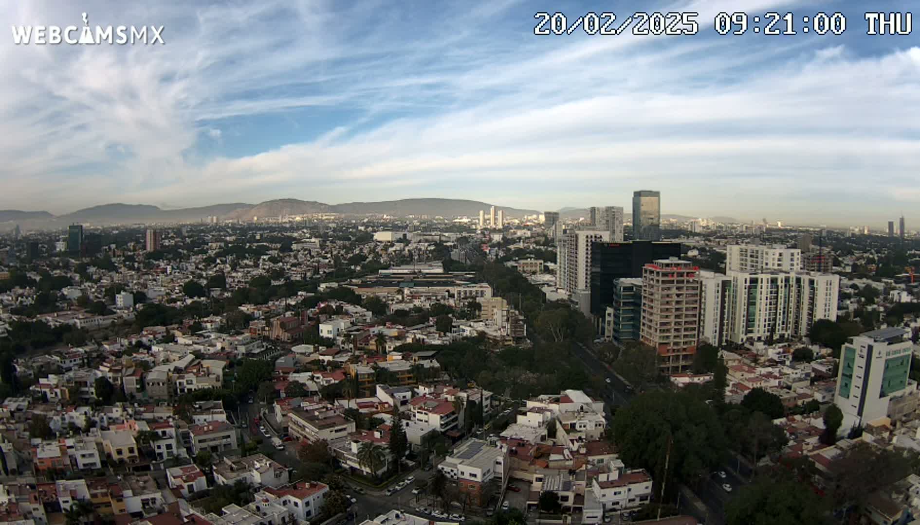 An expansive aerial view of a city under a partly cloudy sky shows numerous residential buildings, taller structures, and mountains in the distance.