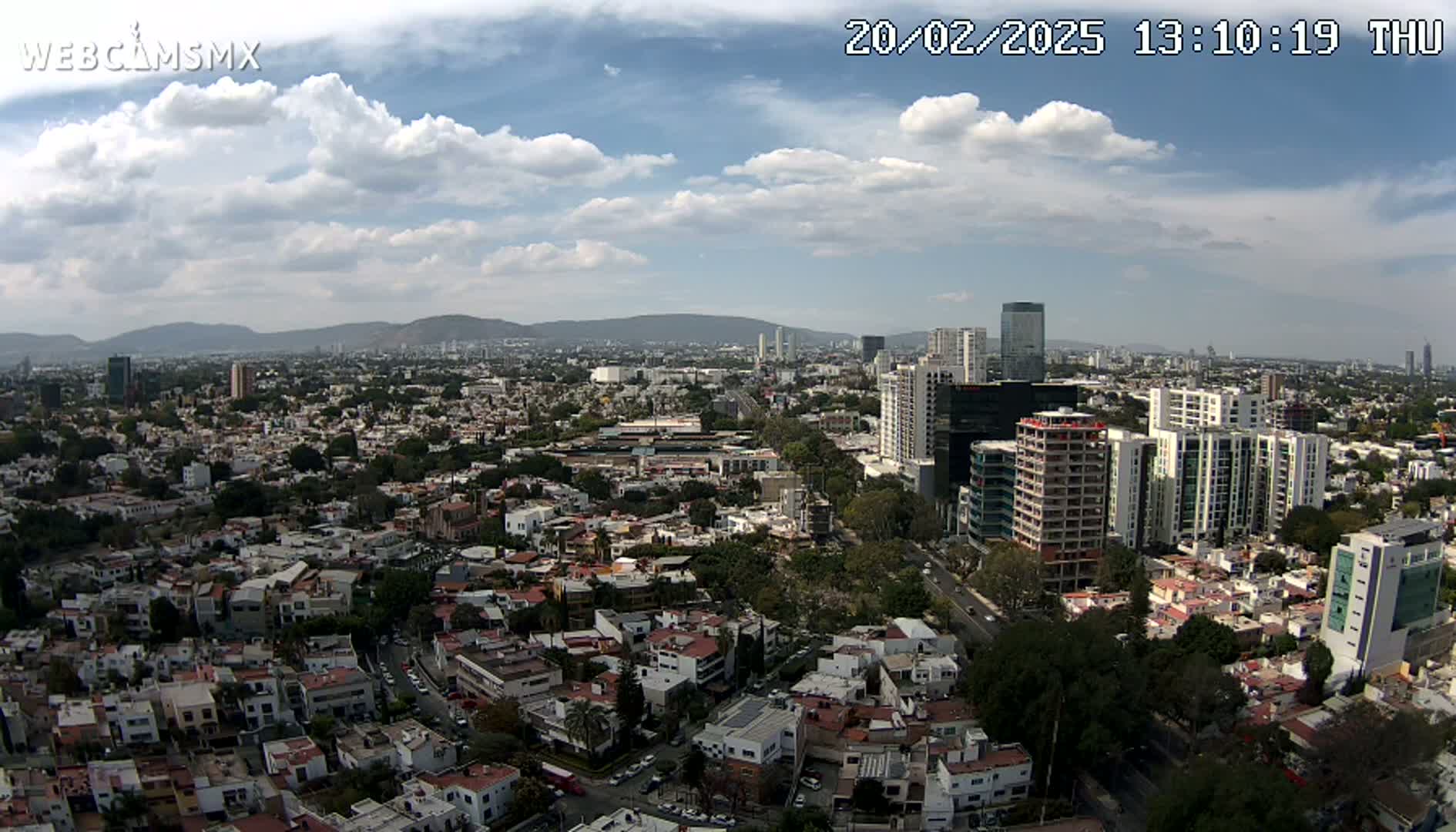 A partly cloudy sky overlooks a sprawling city of low-rise and high-rise buildings, extending to distant mountains.